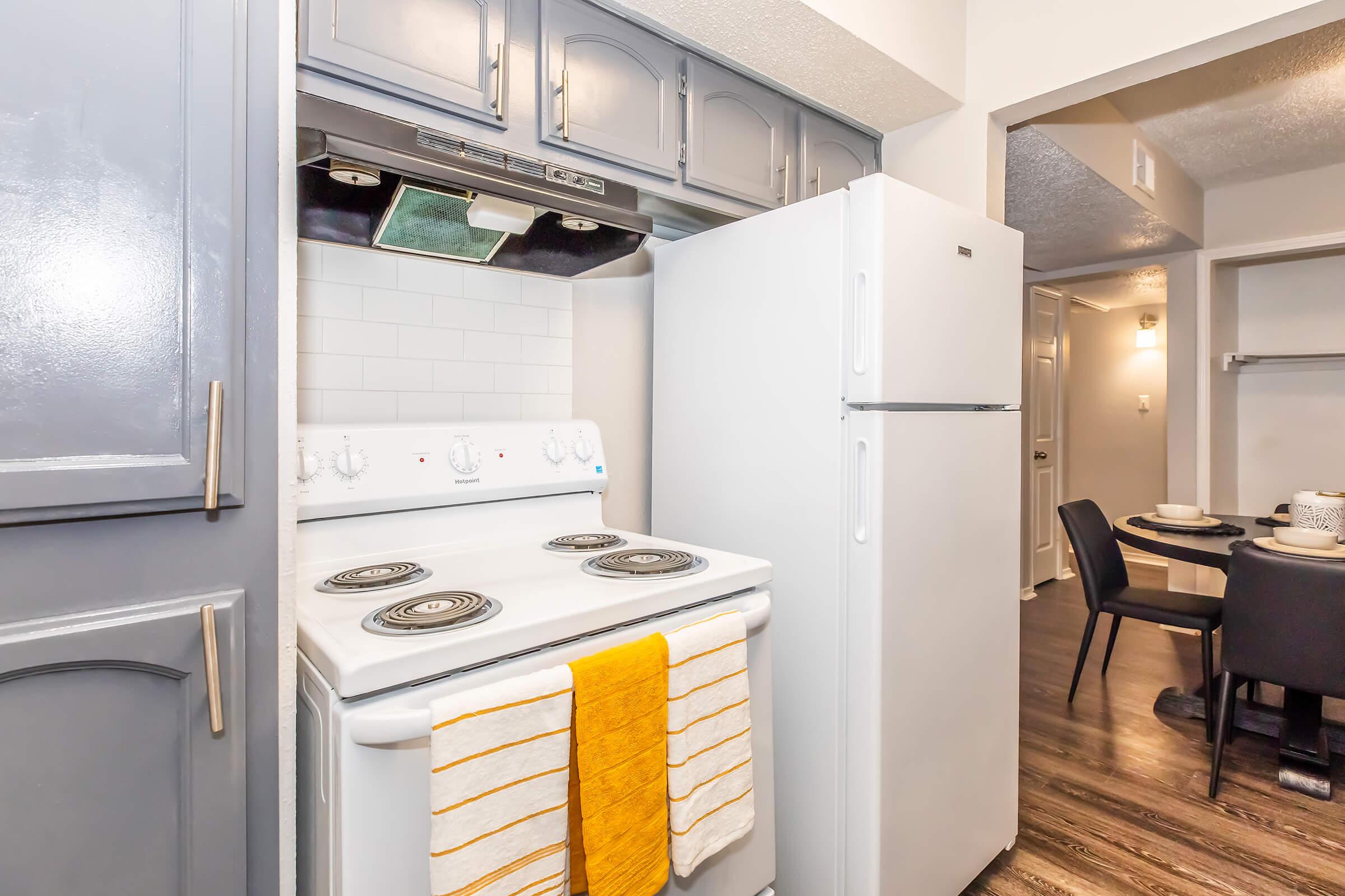 A compact kitchen featuring a white stove with four burners, a white refrigerator, and light gray cabinetry. A yellow kitchen towel hangs from the stove. The flooring is a dark wood laminate, and visible in the background are dining chairs and a doorway, indicating a small dining area beyond the kitchen.