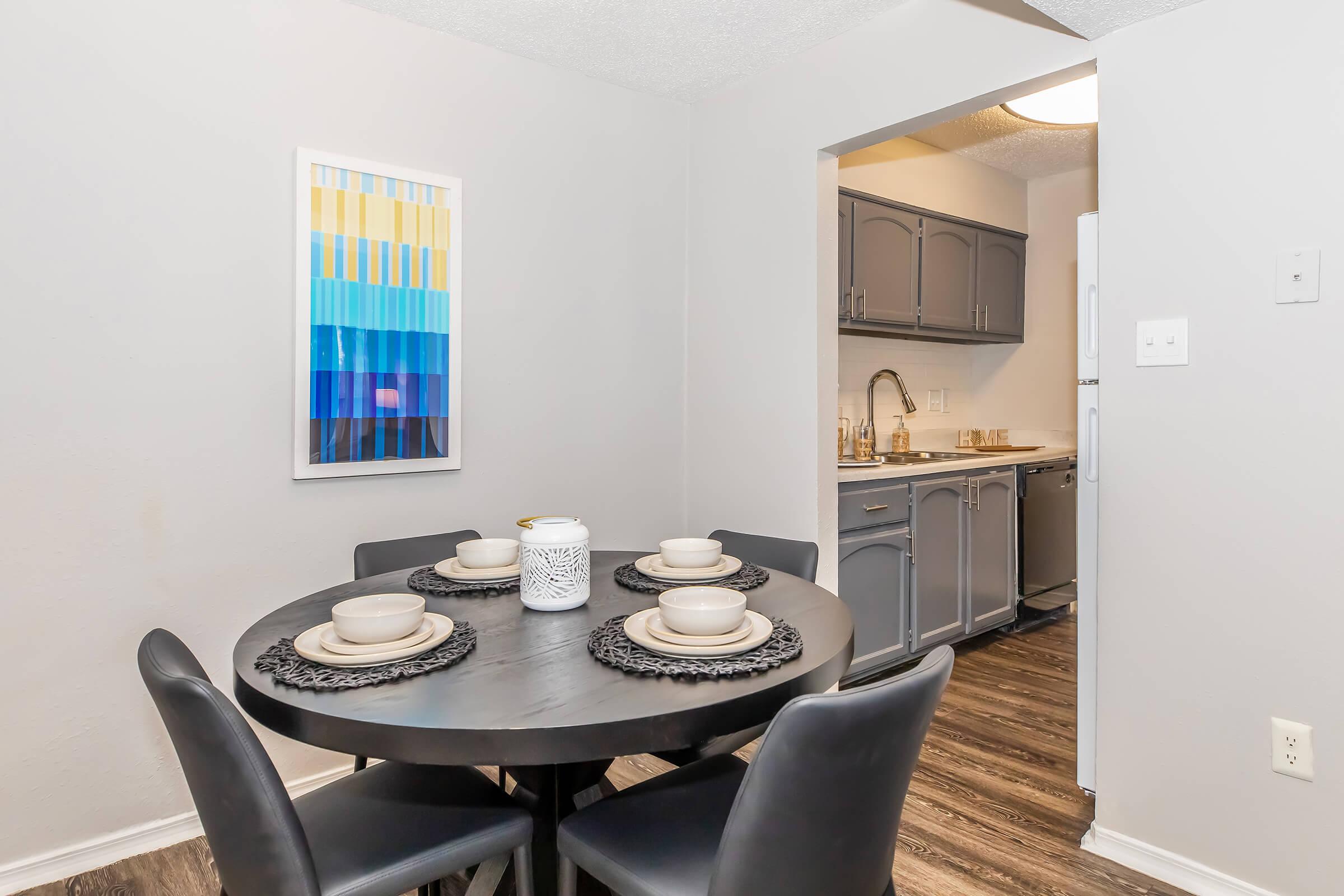 A modern dining area featuring a round black table set with four plates and a decorative centerpiece. Behind the table, a light-colored kitchen is visible, showcasing gray cabinetry and appliances. The wall is adorned with an abstract blue and yellow artwork. The space has a clean and contemporary feel.
