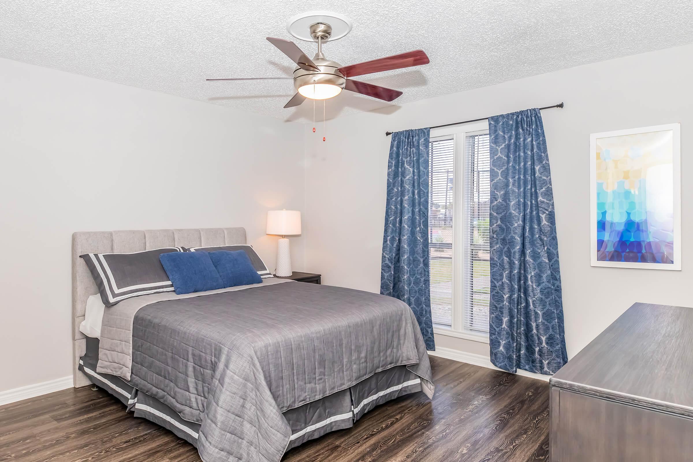 A cozy bedroom featuring a queen-sized bed with gray bedding, a decorative blue accent pillow, a bedside lamp, and a modern ceiling fan. The room has light gray walls, dark wooden flooring, and blue curtains over a window, with abstract wall art nearby.