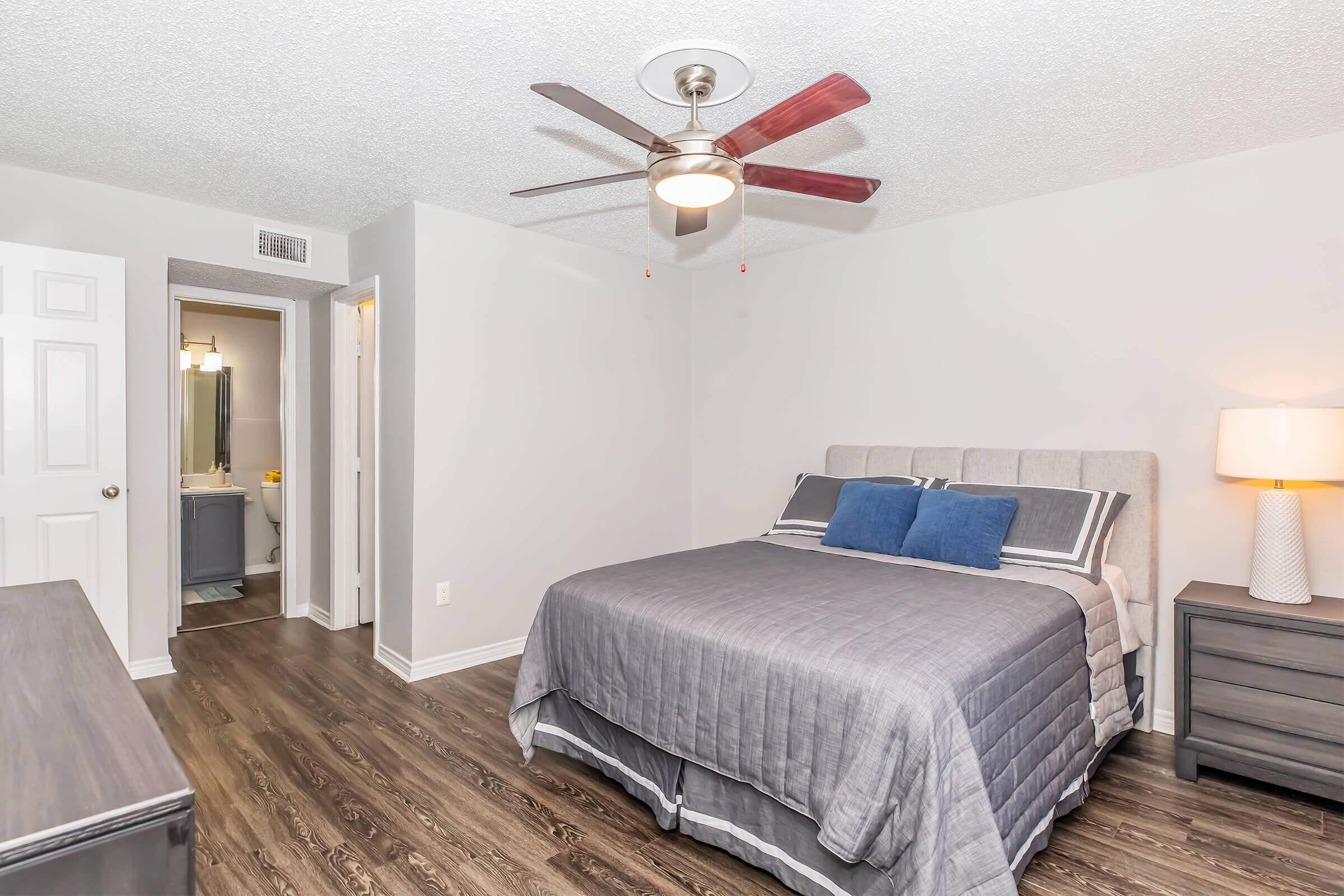 Spacious bedroom featuring a gray upholstered bed with blue accent pillows, a modern ceiling fan, and stylish nightstands. A bathroom doorway is visible in the background, and the room has a light gray color scheme with wooden flooring.