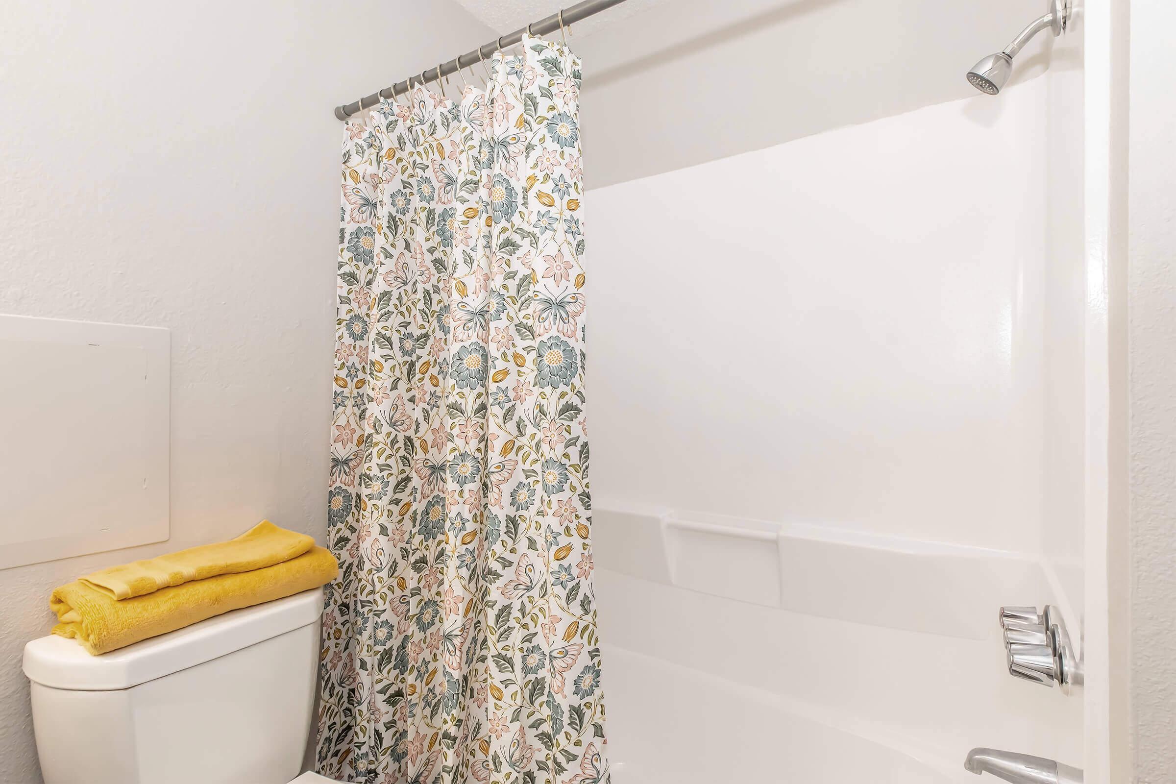 A bathroom scene featuring a shower area with a floral-patterned curtain, a white bathtub, and a toilet. A neatly folded yellow towel rests on top of the toilet tank. The walls are painted light gray, contributing to a clean and modern aesthetic.