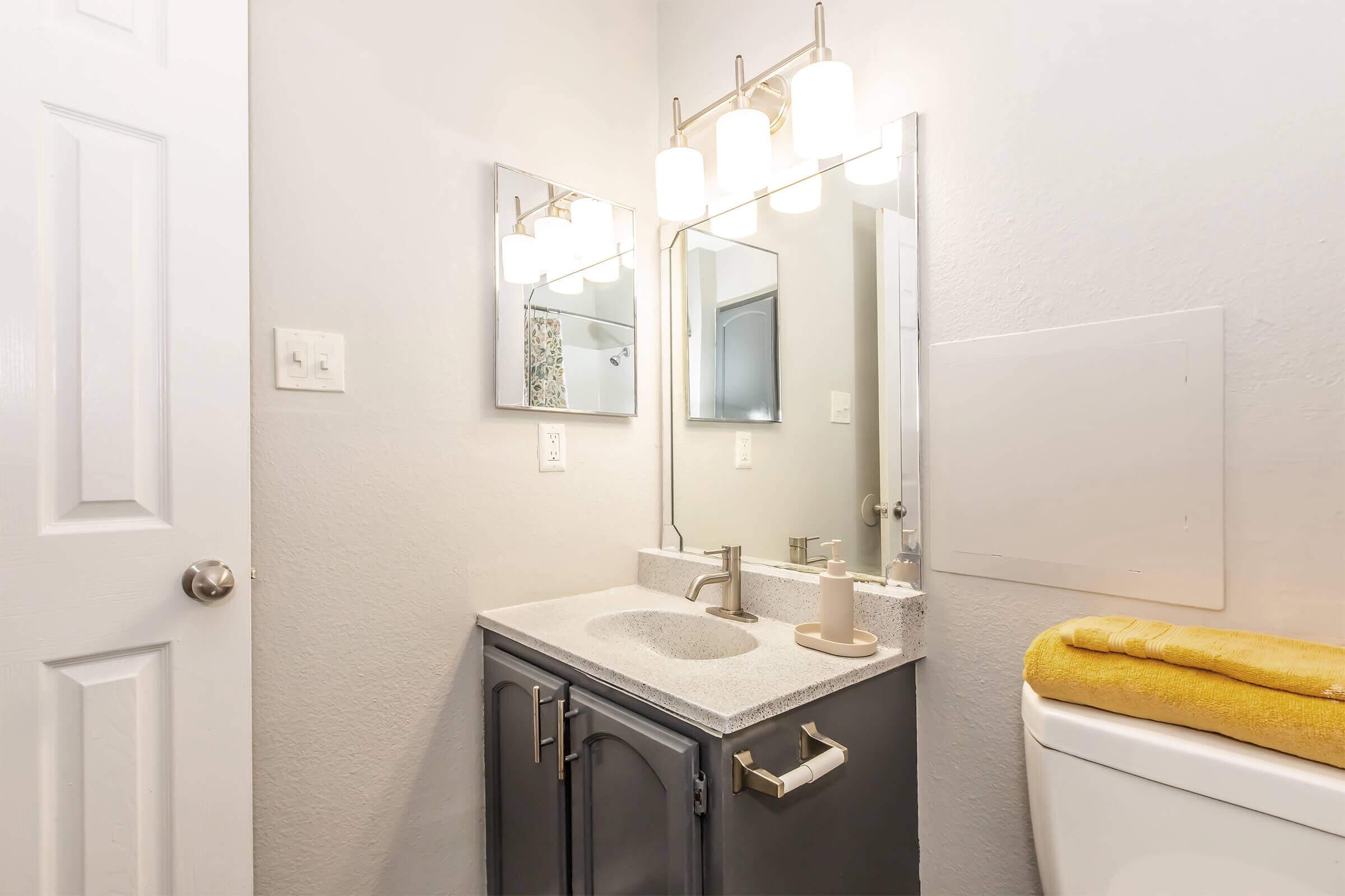 A modern bathroom featuring a grey vanity with a sink, a large mirror with four light fixtures above it, and a yellow towel neatly placed on the toilet. The walls are painted in a light color, and a small cabinet is visible. The overall decor is clean and contemporary.