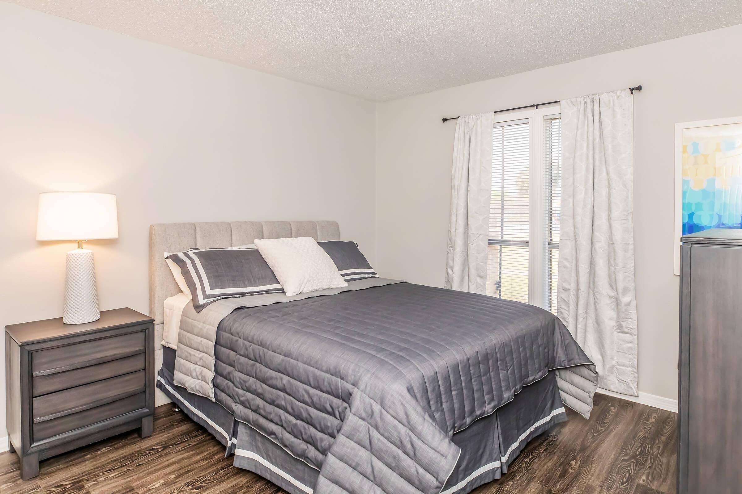 A cozy bedroom featuring a queen-sized bed with gray bedding and decorative pillows, a bedside table with a lamp, and window curtains. The walls are light-colored, and there is a stylish piece of artwork on the wall, creating a calm and inviting atmosphere.