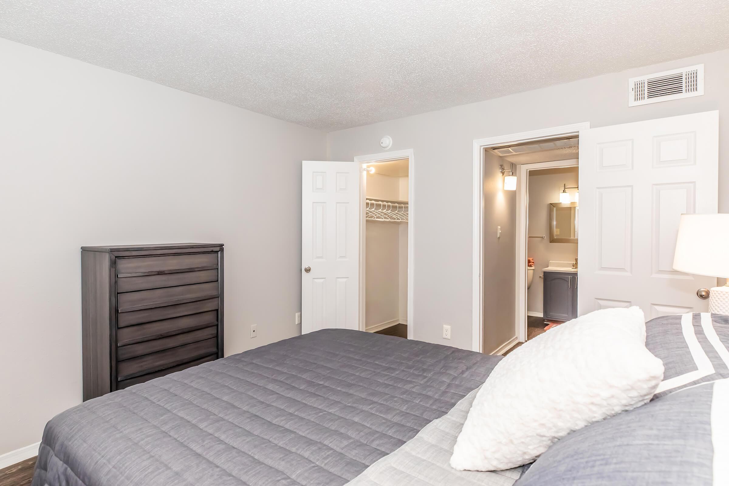 A cozy bedroom featuring a neatly made bed with gray bedding, a wooden dresser, and two closed doors leading to a closet and bathroom. The walls are painted light gray, and the room has a soft ambiance. A bedside lamp adds warmth to the space.