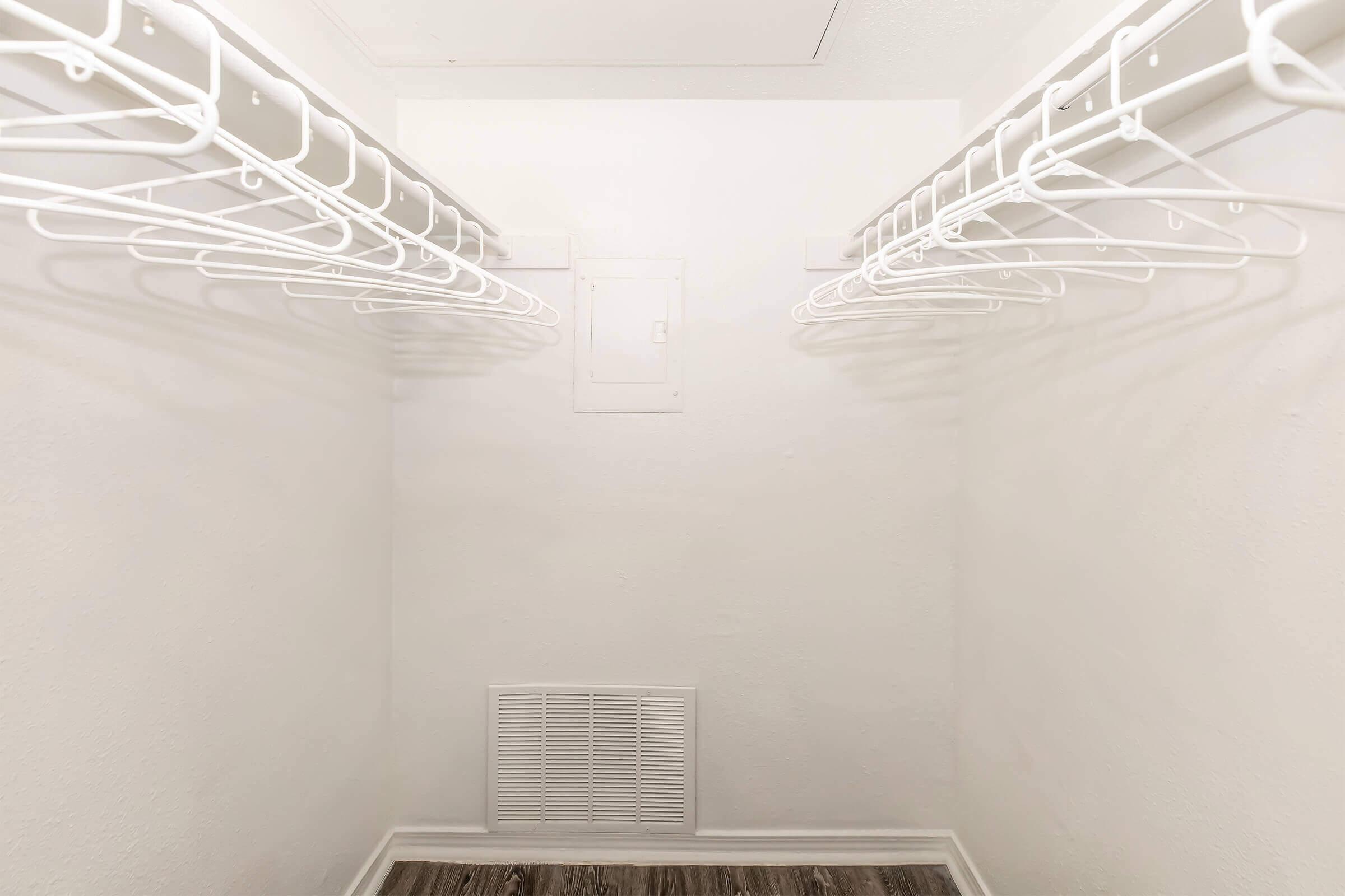 A clean, empty closet featuring white wire shelving on both sides, with a white wall and floor. There is a vent on the lower wall, and a ceiling access panel visible above. The overall space is bright and uncluttered, ideal for storing clothing or other items.