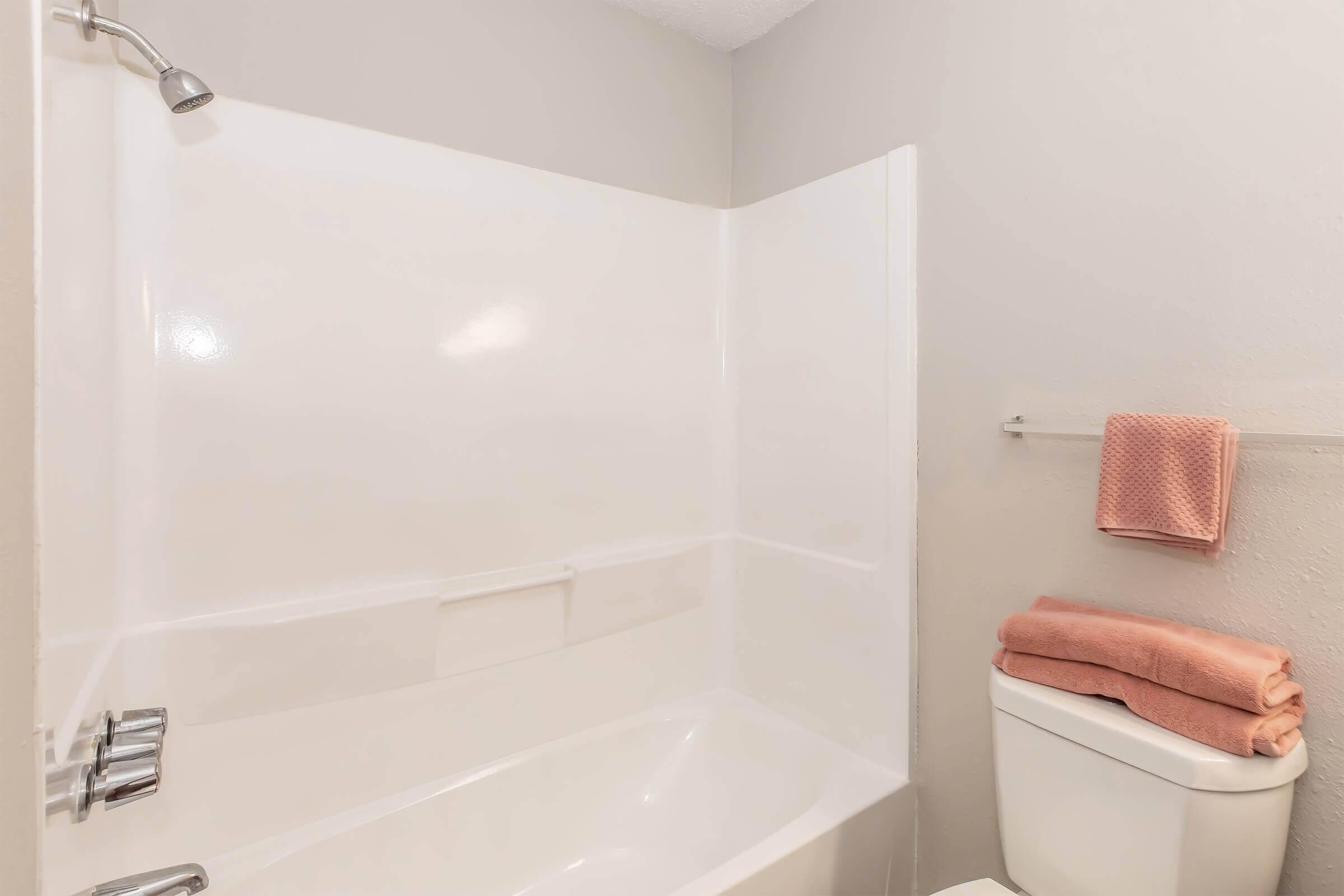 Clean and simple bathroom featuring a white bathtub and shower combination, a modern toilet, and neatly folded pink towels on a rack, with light gray walls providing a neutral backdrop. The overall aesthetic is bright and tidy, ideal for a minimalist design.