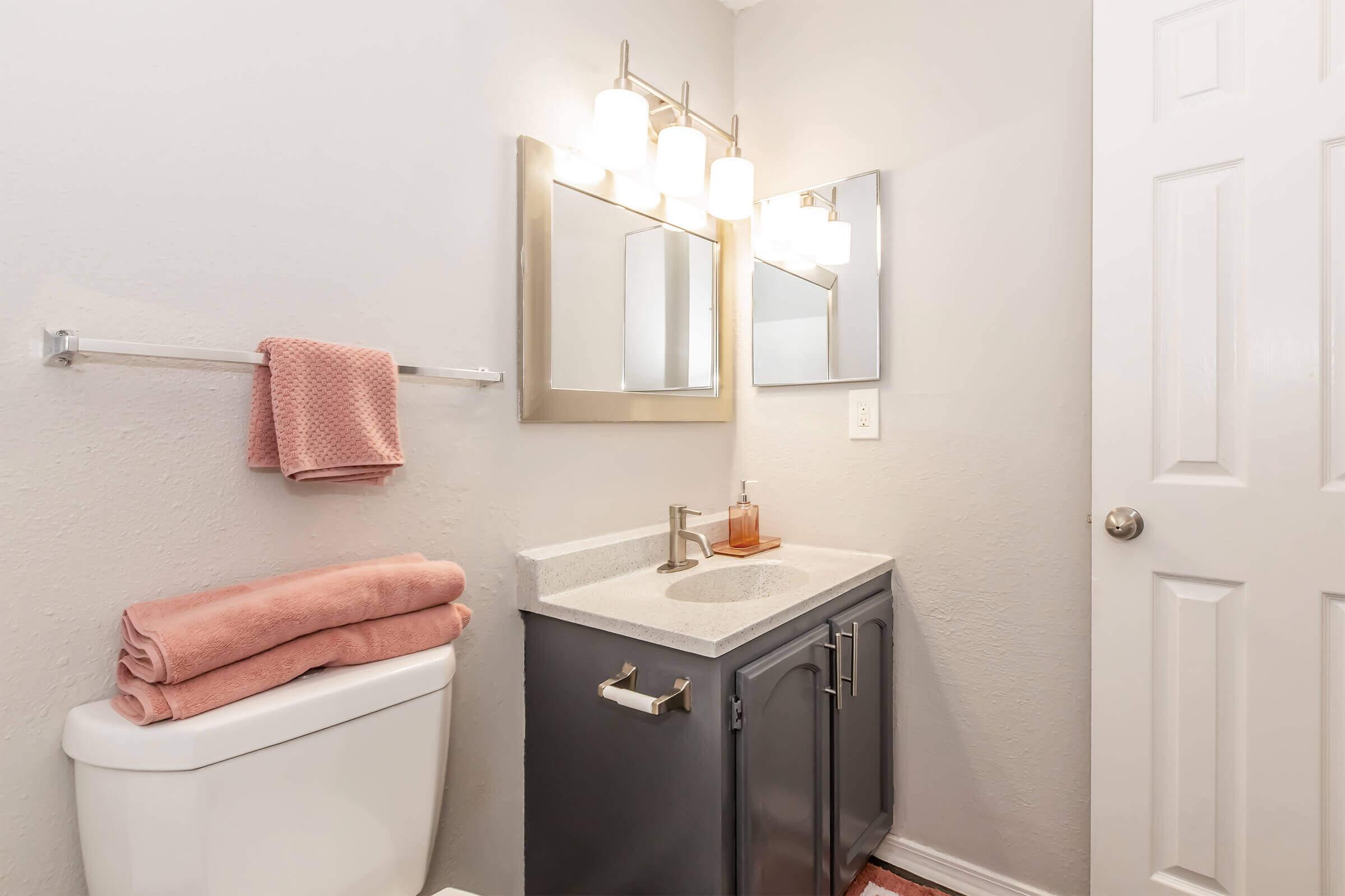 A modern bathroom featuring a gray wall, a sleek vanity with a white countertop, a large mirror, and stylish light fixtures. A toilet is visible, along with neatly folded pink towels and a pink washcloth hanging on a towel rack. The overall aesthetic is clean and contemporary.