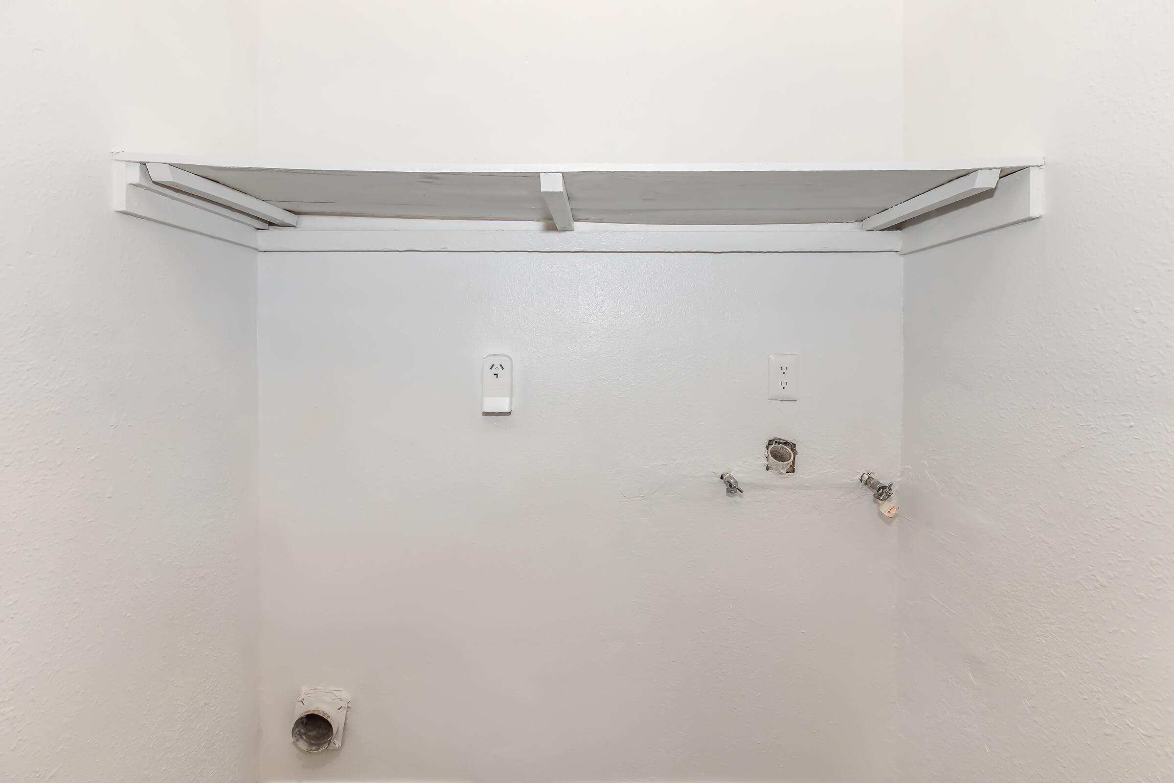 A bare wall with a white shelf mounted above, featuring electrical outlets and plumbing connections visible. The wall is smooth and light-colored, emphasizing the simplicity of the space.