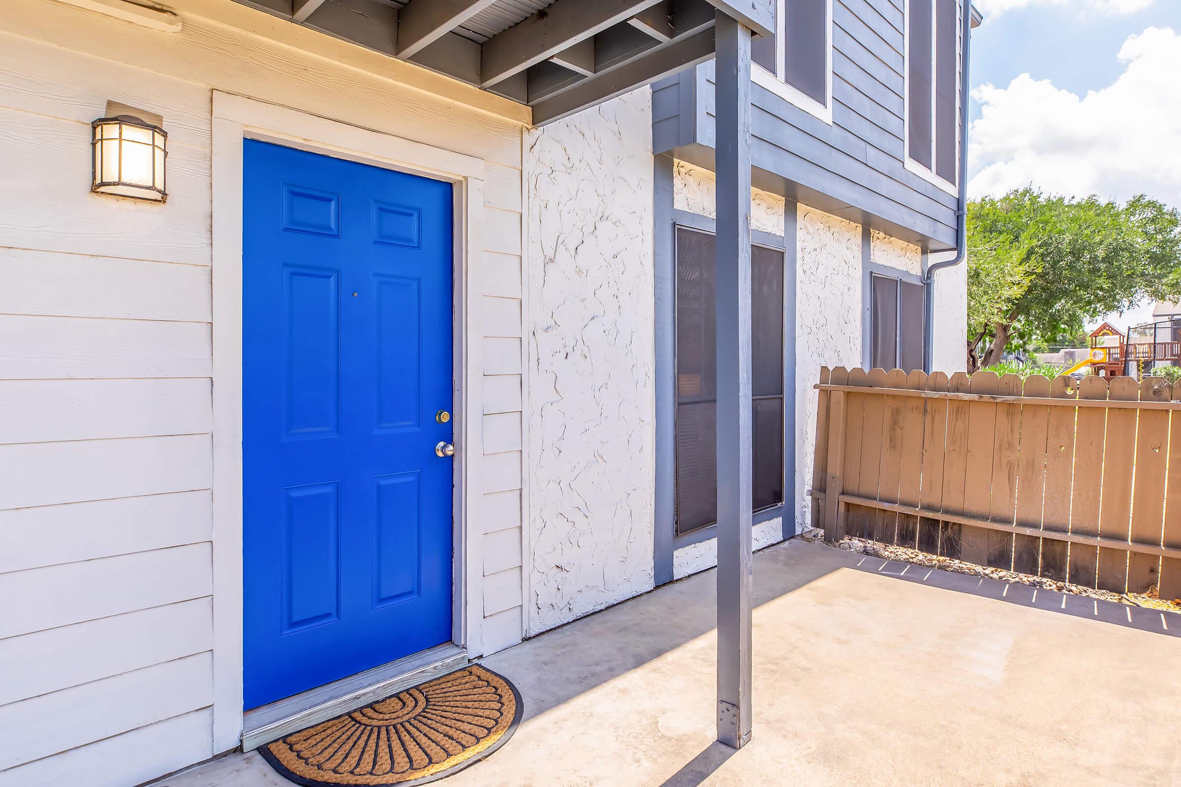 A bright blue front door is framed by a beige exterior wall, complemented by a small round welcome mat at the doorstep. A covered entryway features a light fixture, and a wooden fence is visible to the right, suggesting an outdoor space with greenery beyond.