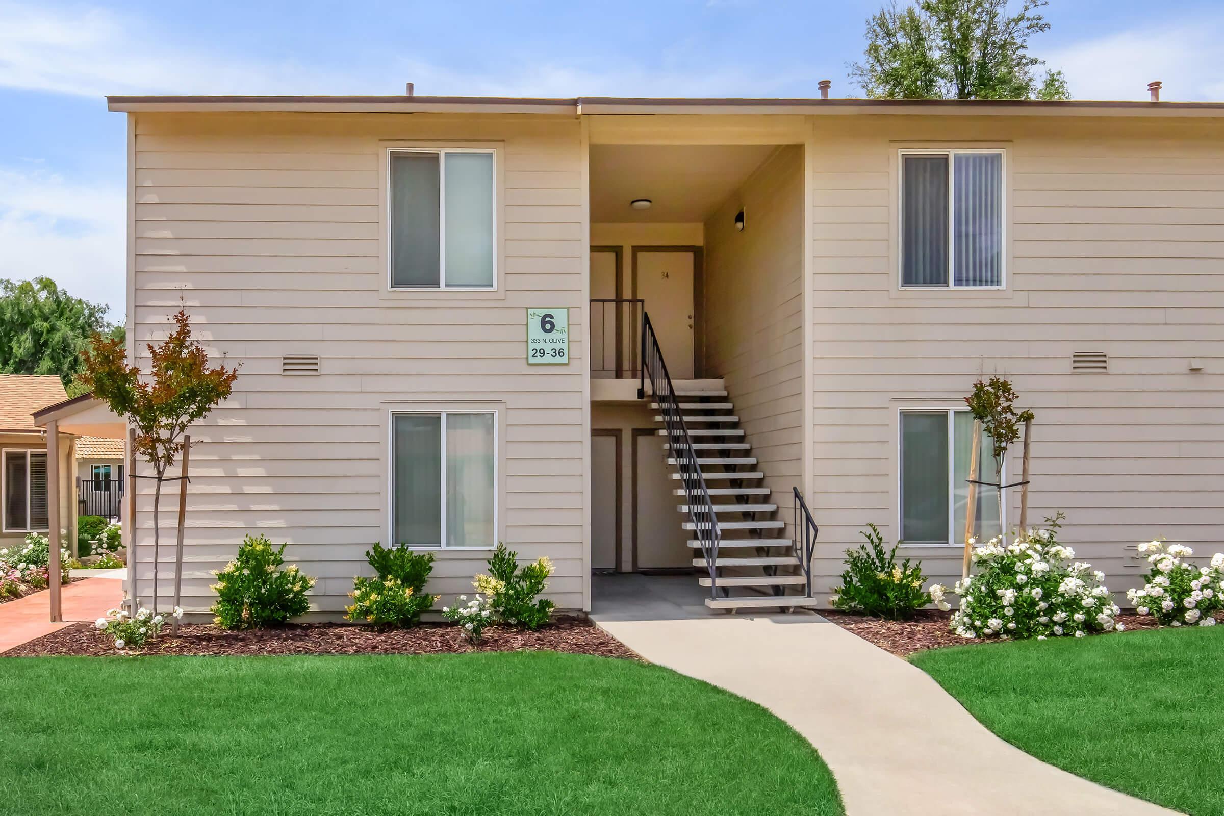 Two-story apartment building featuring a light-colored exterior with a front entrance and a staircase leading to the second floor. Lush green grass and flowering shrubs surround the pathway leading to the door. A sign indicates the unit number. Bright, clear sky in the background.