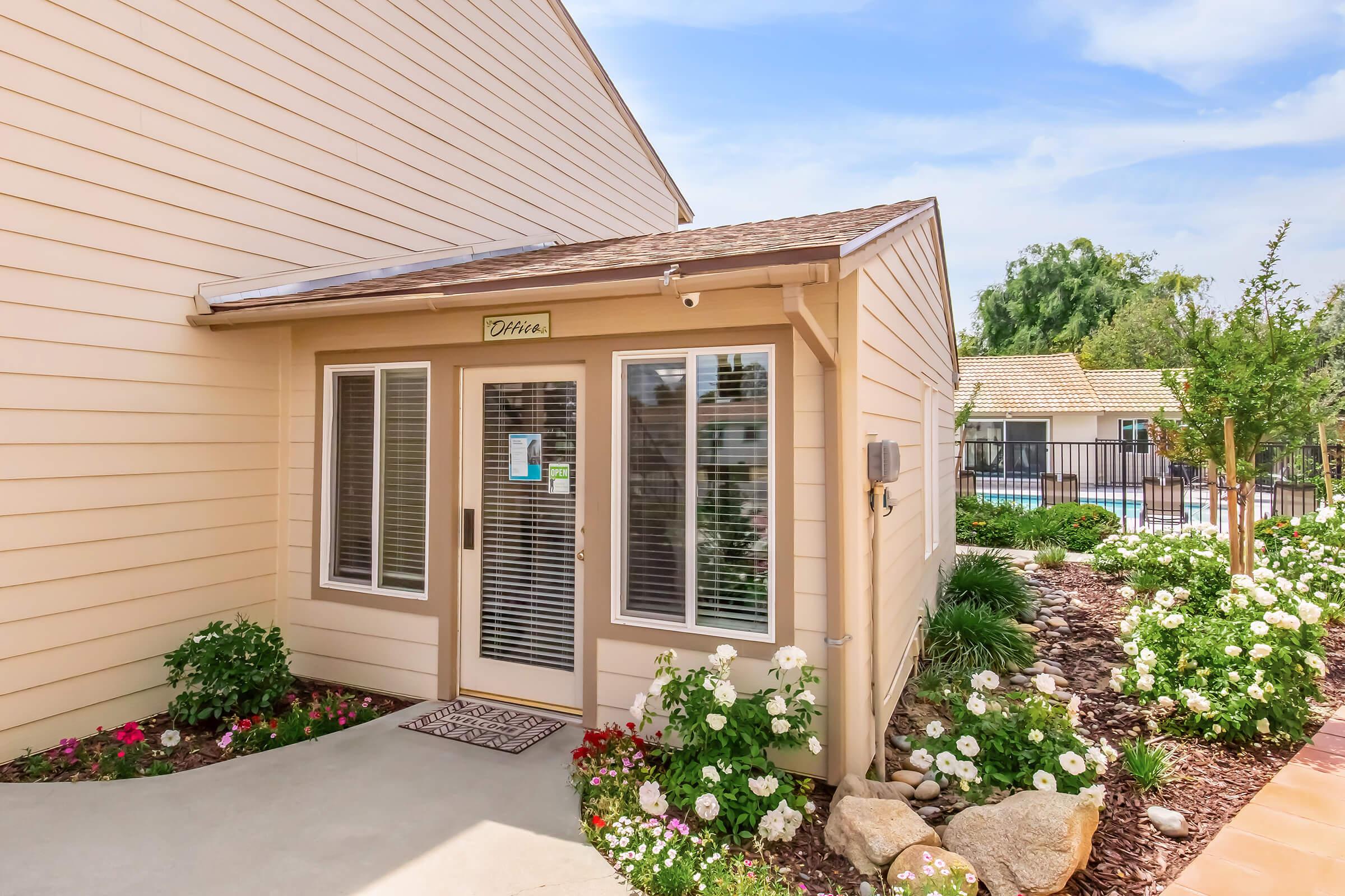 A cozy entrance to a light-colored house featuring a welcoming front door with large windows, surrounded by lush green plants and colorful flowers. A well-maintained pathway leads to the entrance, set against a backdrop of a clear blue sky and scenic landscaping.