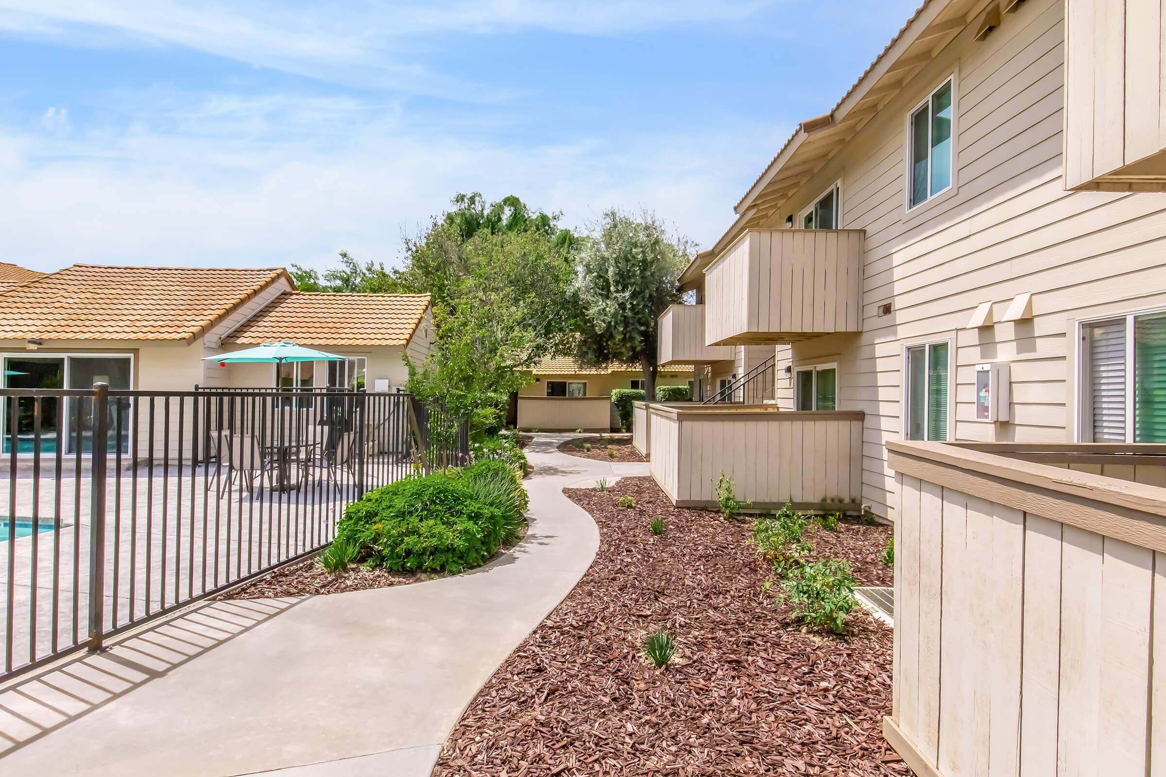 Pathway winding through a landscaped area with shrubs and mulch, leading to a two-story apartment building. On the left, a pool area is enclosed by a fence, with a sun umbrella visible. The scene features bright blue skies and greenery, creating a relaxed outdoor atmosphere.