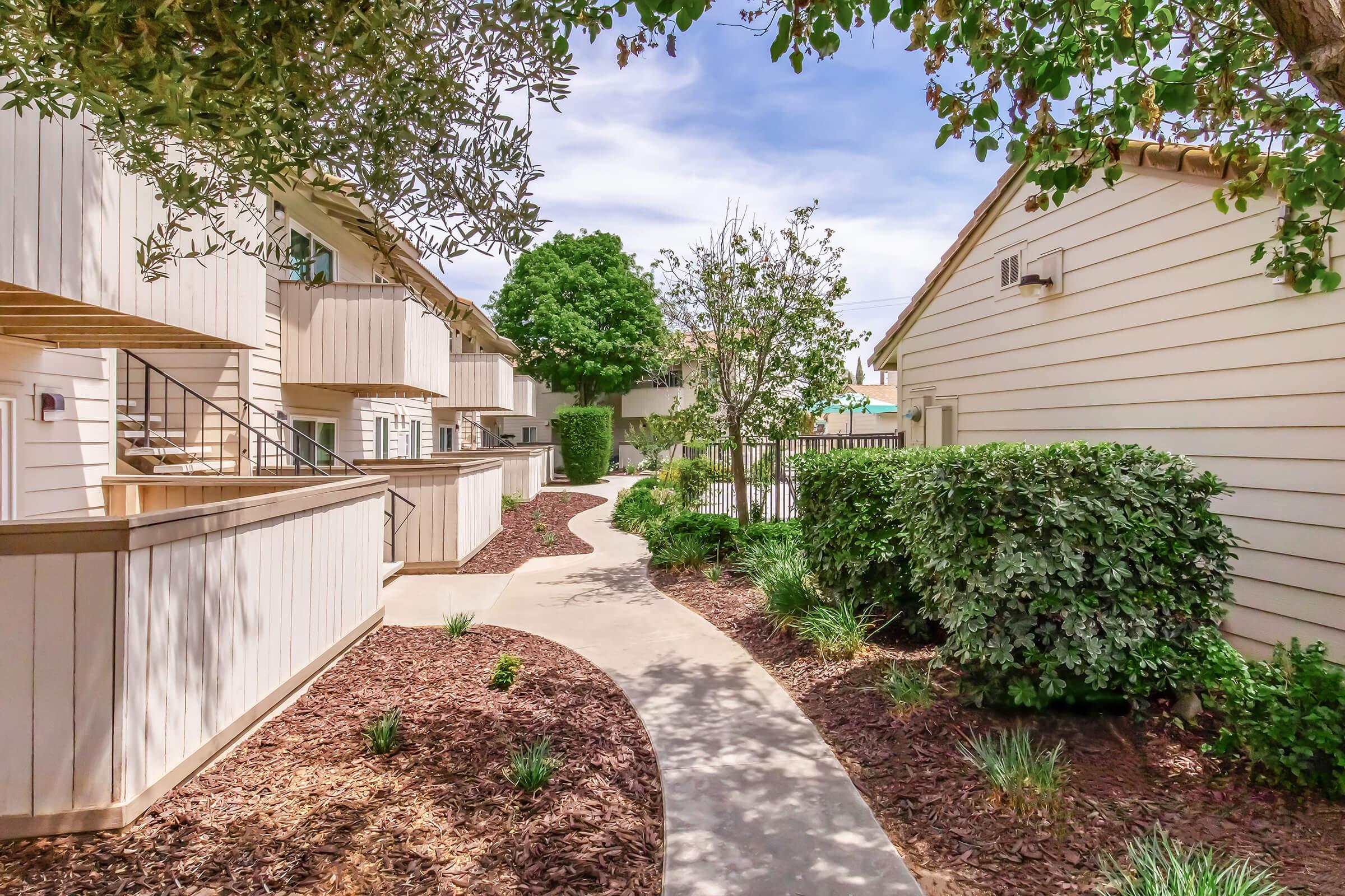 A well-maintained pathway meanders through a landscaped area with neatly trimmed bushes and trees, flanked by light-colored buildings. The setting is bright and open, suggesting a pleasant residential environment.