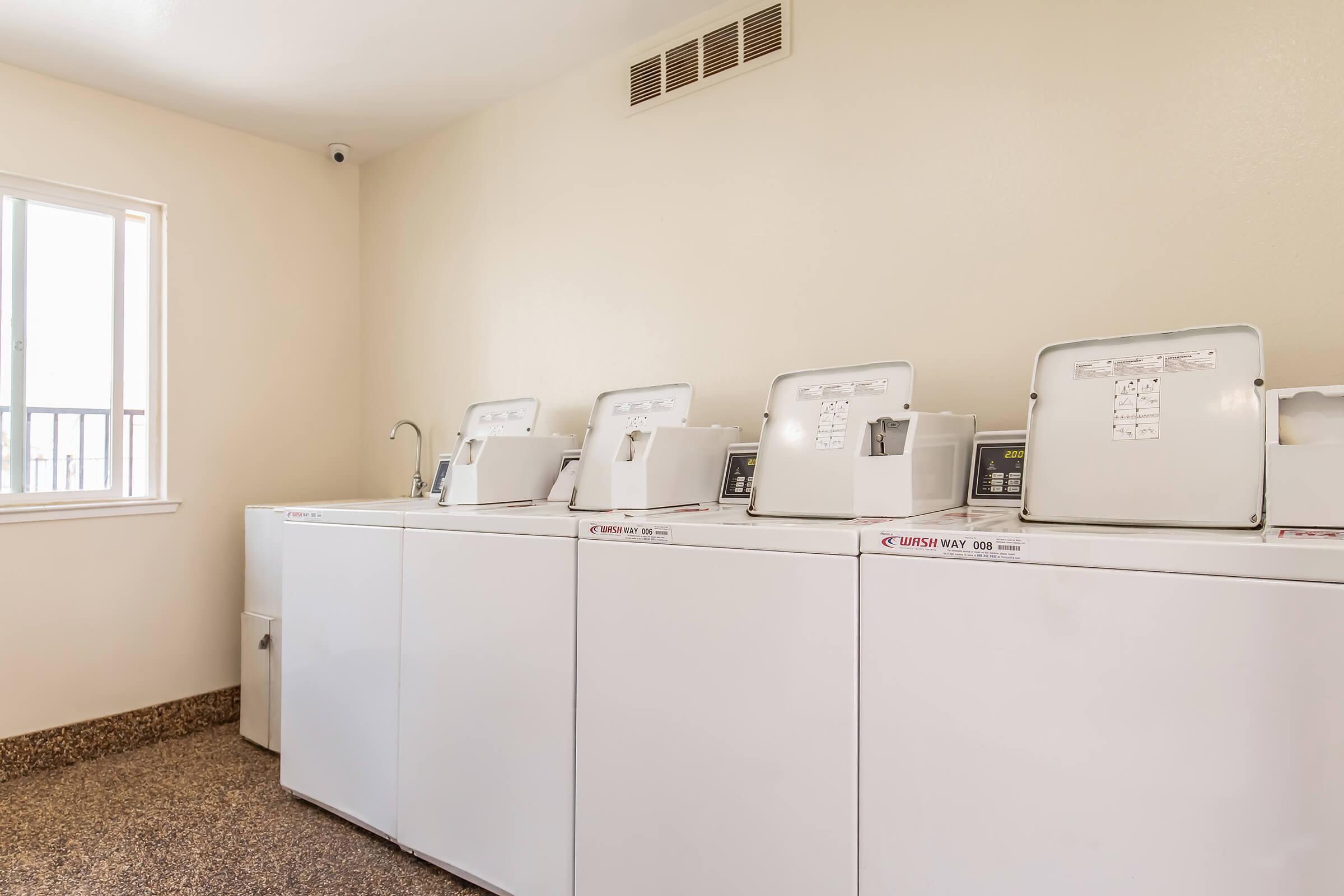 A clean laundry room featuring four white washing machines with visible control panels. The space is well-lit with a window providing natural light, and the flooring is a dark textured surface. A small sink is also present in the area.