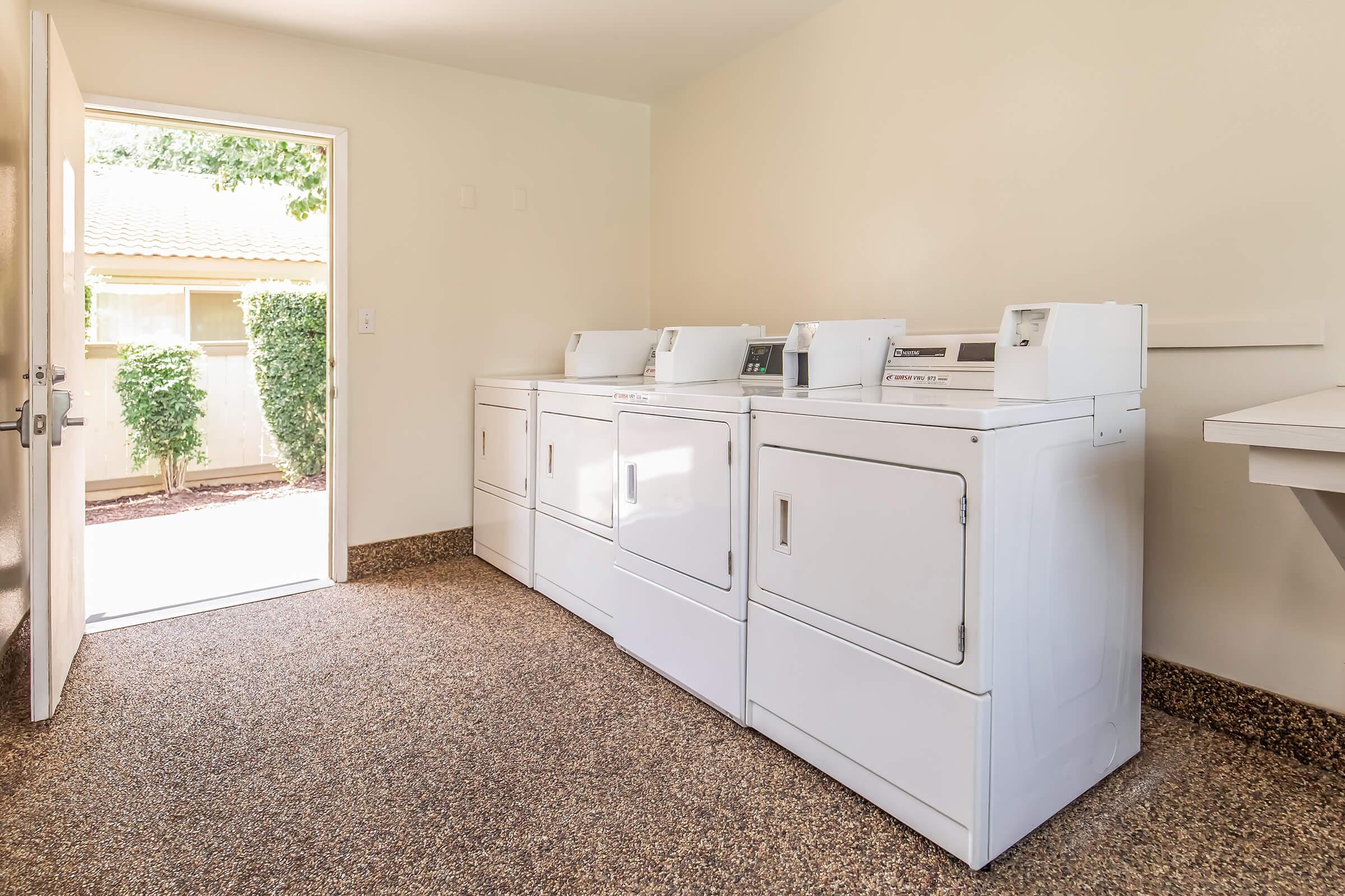 A clean laundry room featuring three white washing machines and dryers on a textured floor. There is a door to the left leading outside, and bright natural light coming in. The walls are a light color, enhancing the room's spacious feel, with a small window for additional light.