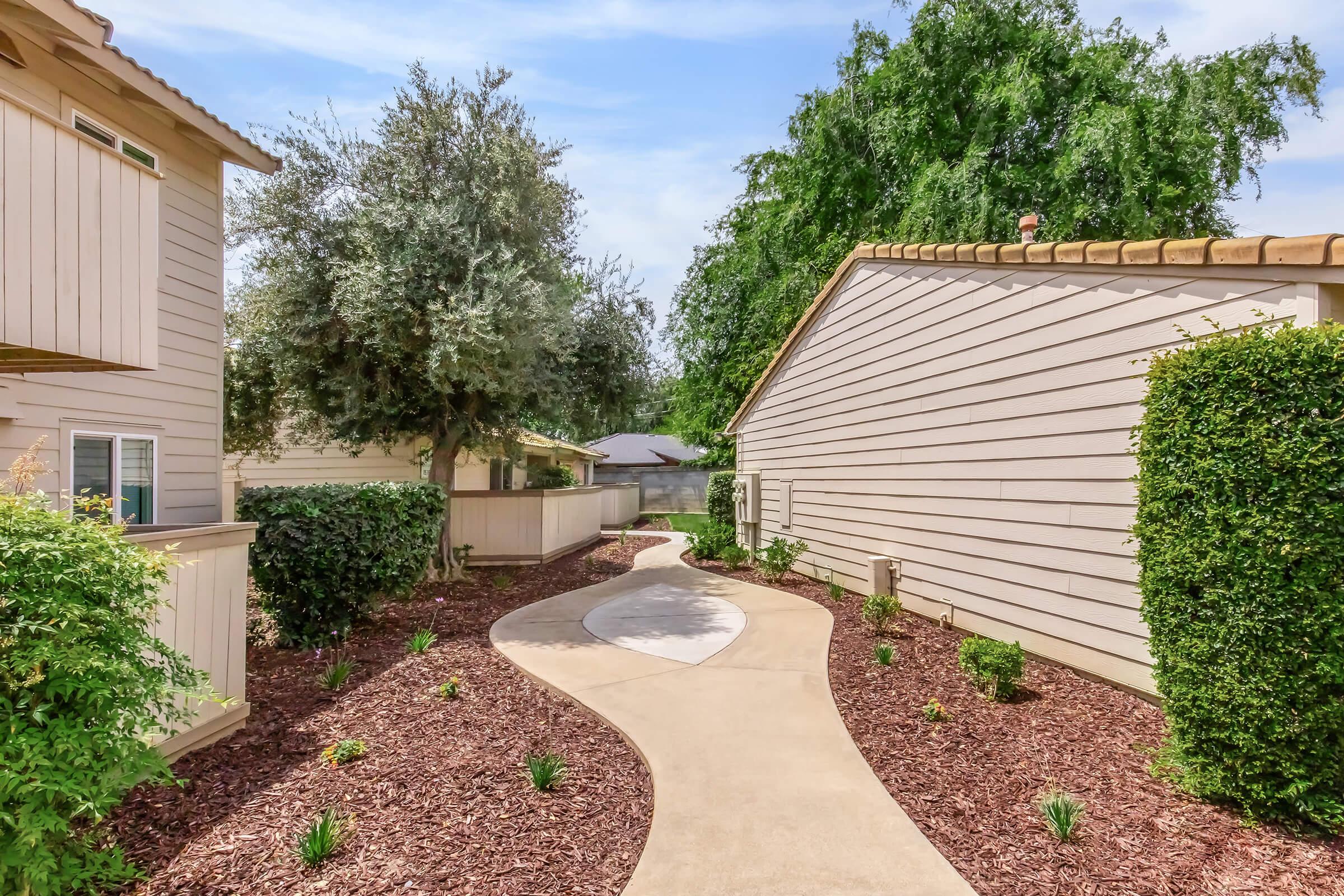 A landscaped path winding between two buildings, lined with low shrubs and mulch. Lush greenery and trees are visible on either side, creating a serene outdoor setting. The pathway is smoothly paved, leading to a distant area with more greenery. The sky is clear, indicating pleasant weather.