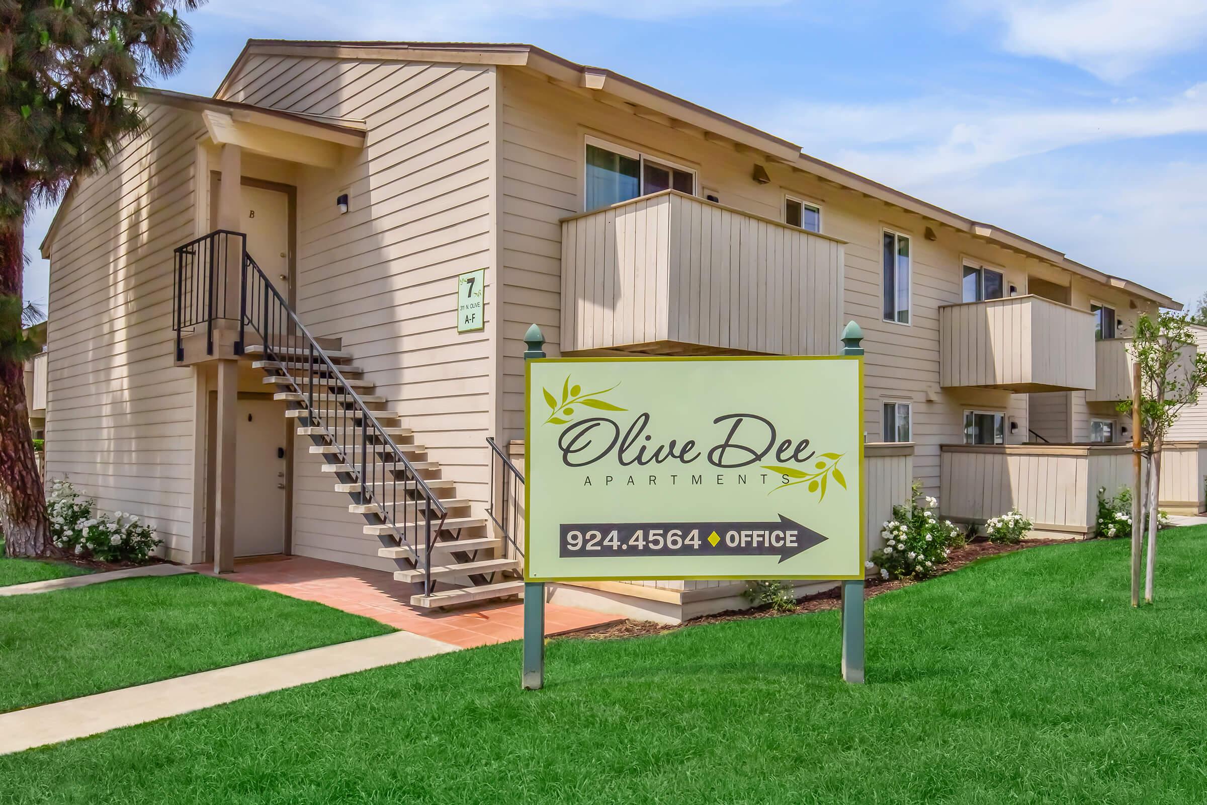 A view of Olive Dee Apartments featuring a sign that displays the name and office phone number. The building is a two-story structure with a staircase, surrounded by well-maintained green grass and shrubbery. There are balconies on the upper level, and the sky is clear with a few clouds.