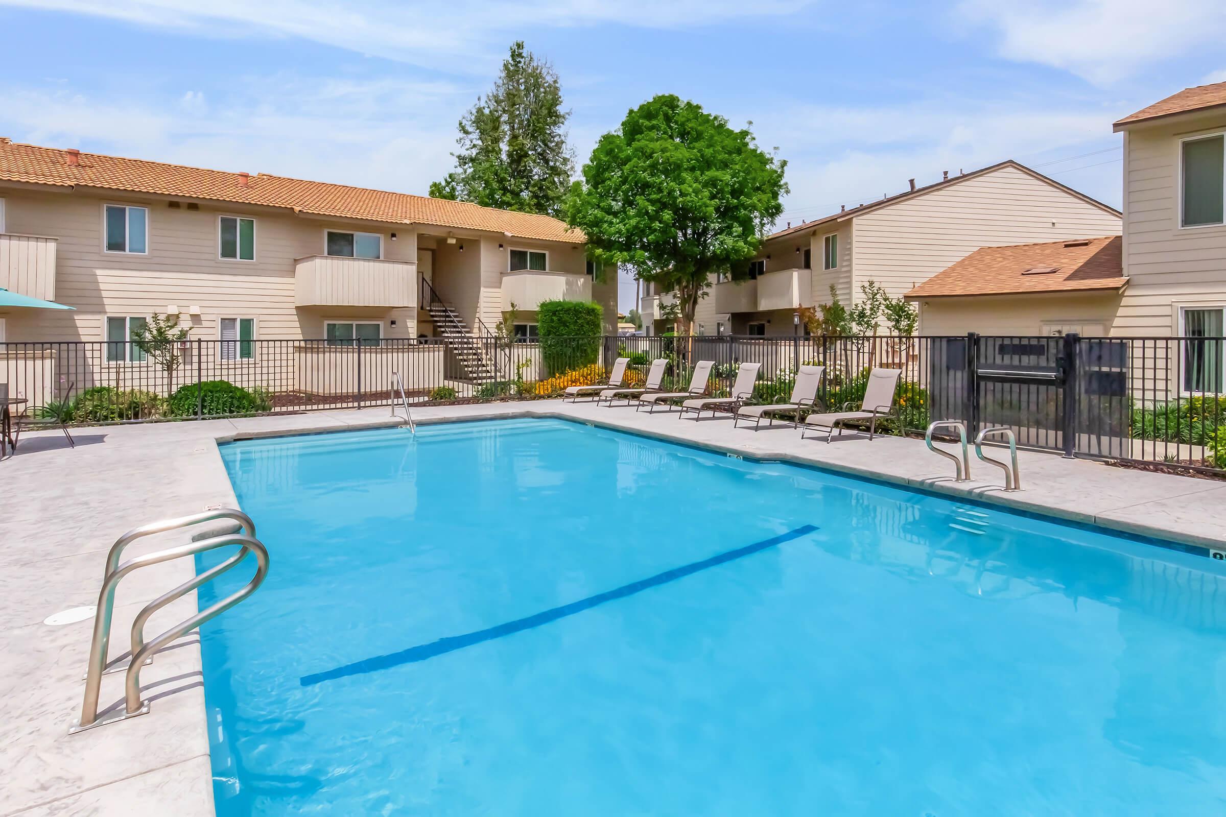 A clear blue swimming pool surrounded by lounge chairs and a fenced area, with two multi-story apartment buildings in the background. Lush greenery and trees add to the pleasant outdoor setting, and the sky is bright with a few clouds.