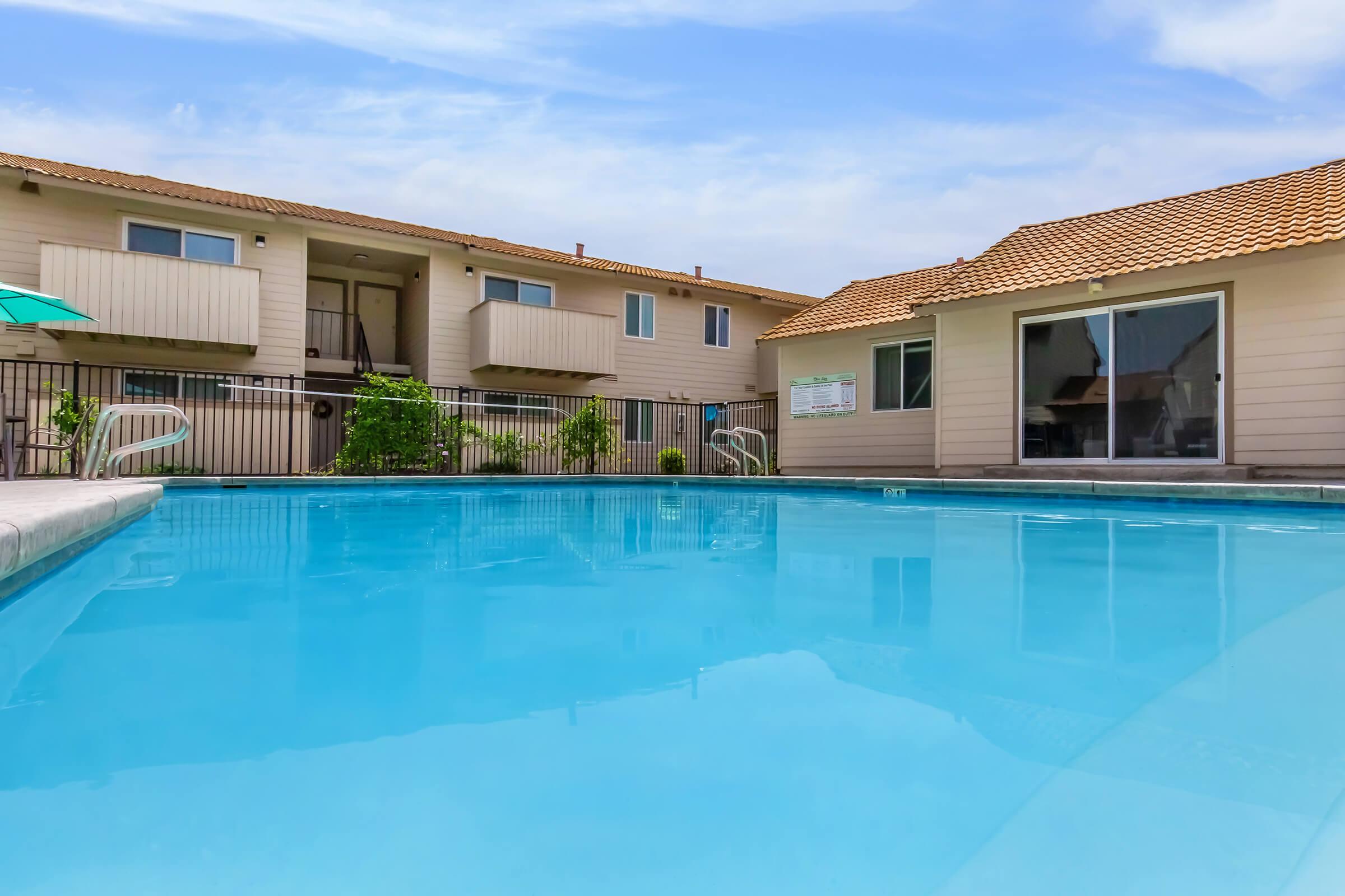A clear blue swimming pool surrounded by apartment buildings with balconies and a tiled roof. Lush green plants are visible near the pool area, and the sky is bright with a few clouds. The scene conveys a relaxing outdoor atmosphere.