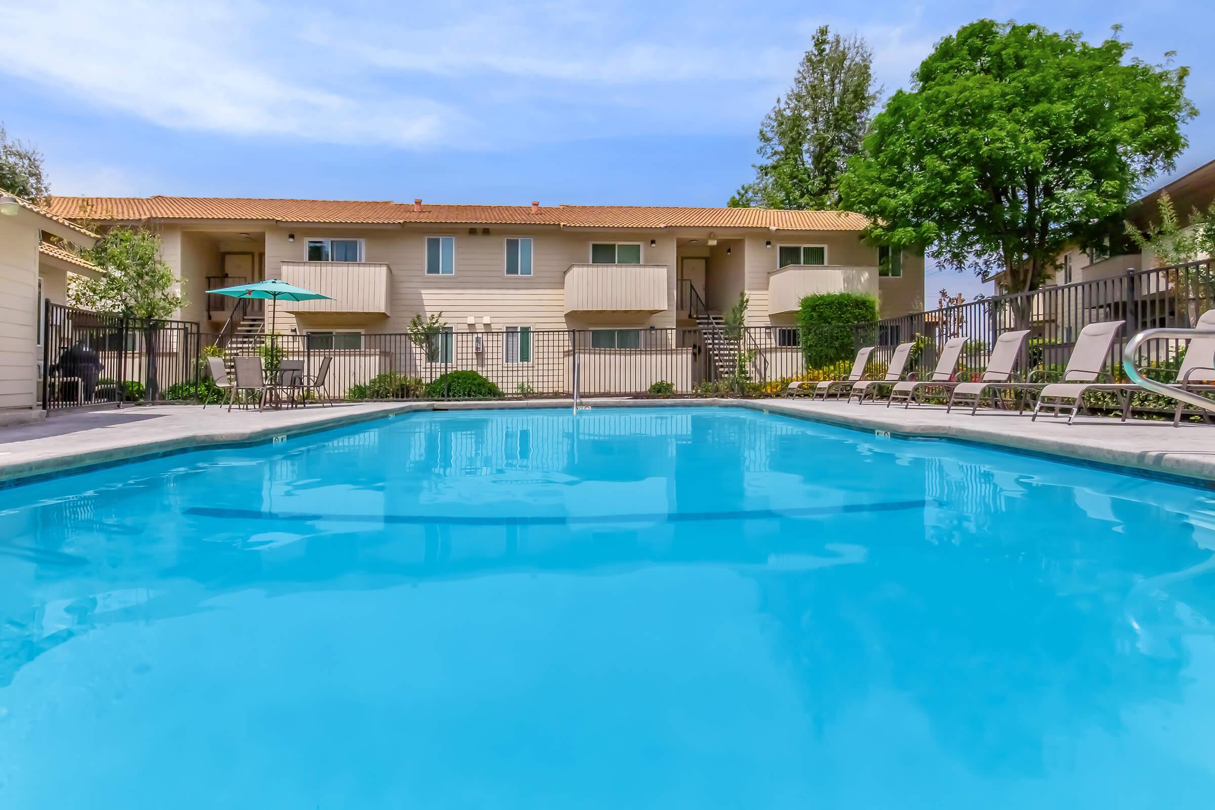 A clear blue swimming pool surrounded by lounge chairs and shaded by trees, with two-story apartment buildings in the background. The area features well-maintained landscaping, providing a serene and inviting atmosphere for relaxation.