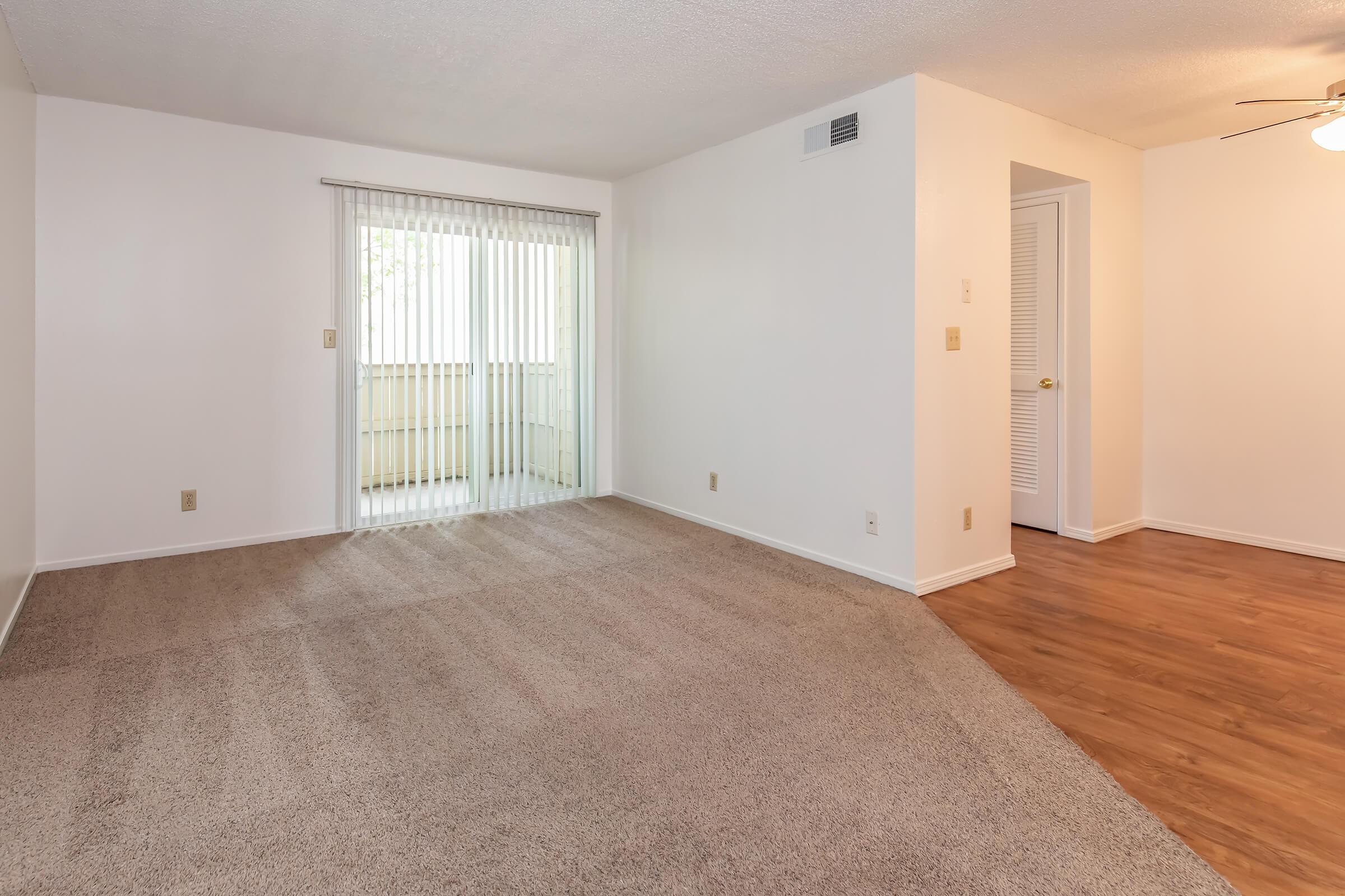 A spacious, empty living room featuring beige carpet flooring, white walls, and a large window with vertical blinds, allowing natural light to enter. The room includes a doorway leading to another area and a ceiling fan above.