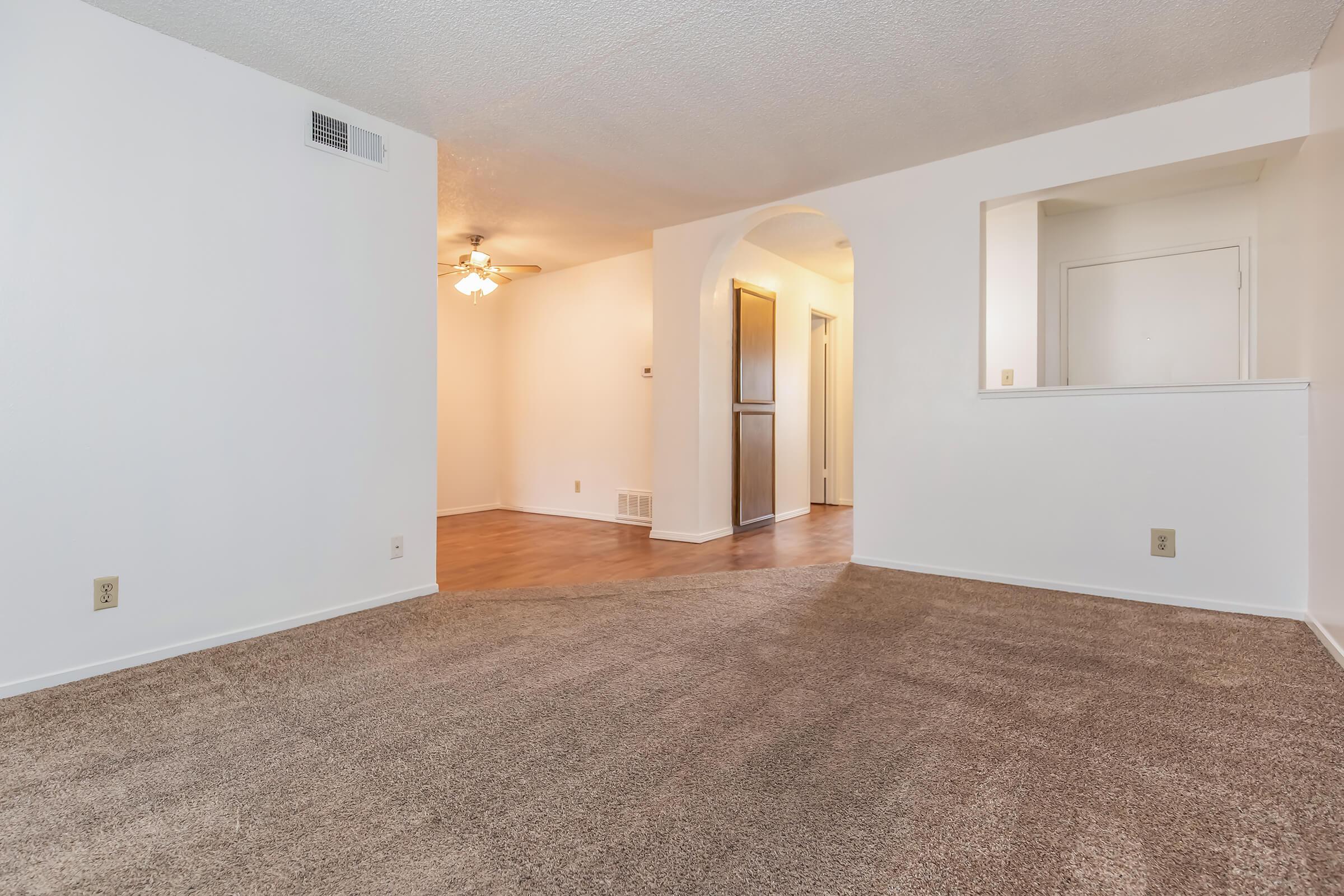 A spacious and well-lit living area featuring light-colored walls, a ceiling fan, and plush carpet flooring. An archway leads to an adjacent room with a contrasting floor and a small window. The layout creates an inviting atmosphere in a modern apartment setting.