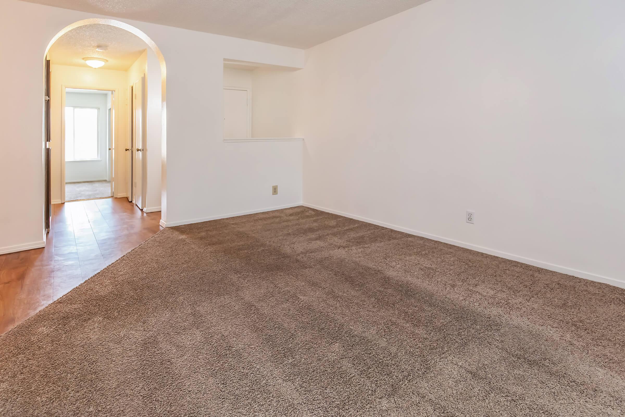 A spacious, empty room with beige carpet and white walls. A doorway leads to a hallway with a light fixture and a window at the end, suggesting natural light. The overall ambiance is bright and neutral, suitable for various decor styles.