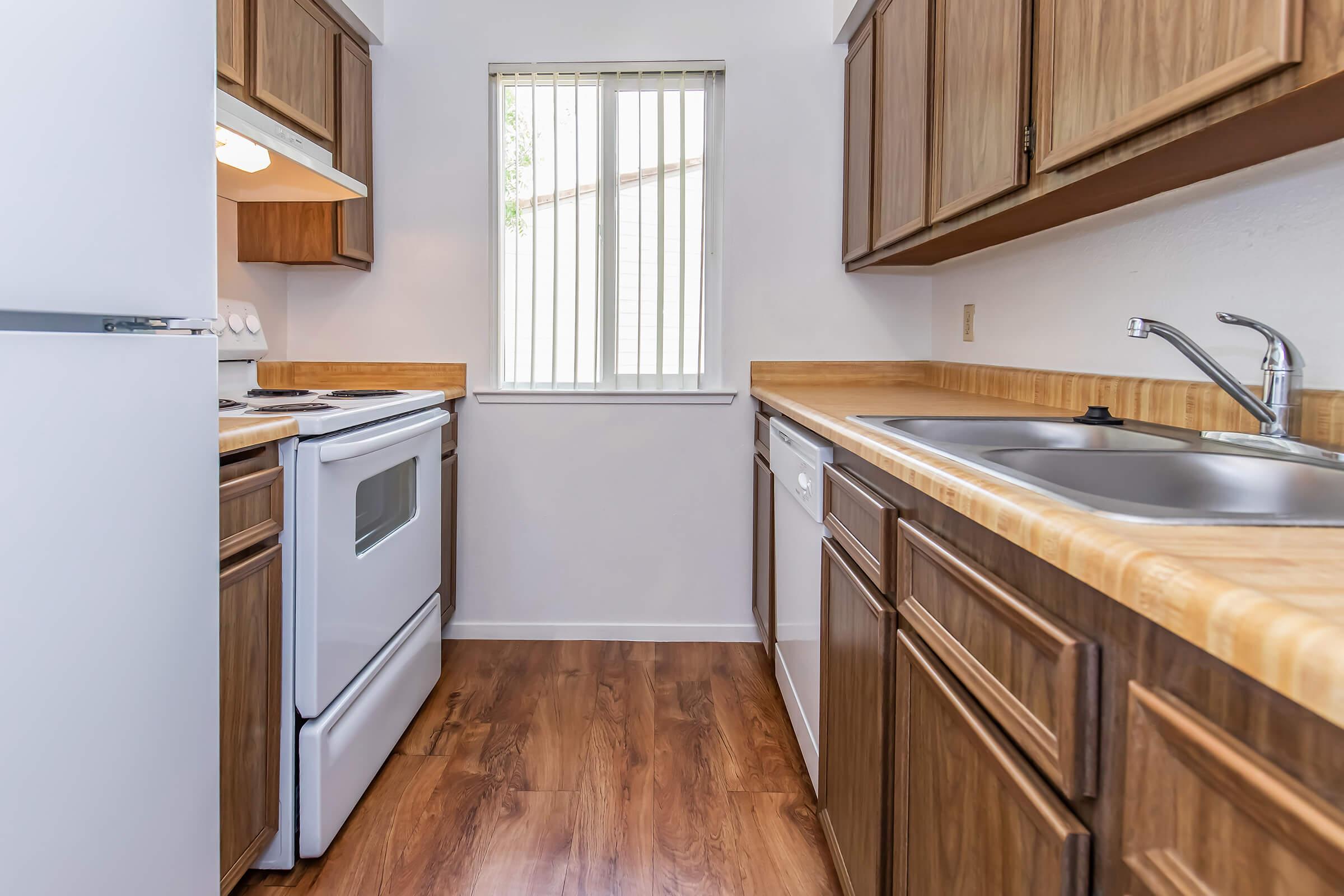 A compact kitchen featuring wooden cabinets, a white stove and refrigerator, a sink, and a dishwasher. The kitchen has a window with vertical blinds, allowing natural light to enter, and a warm, inviting atmosphere with wood-style flooring.