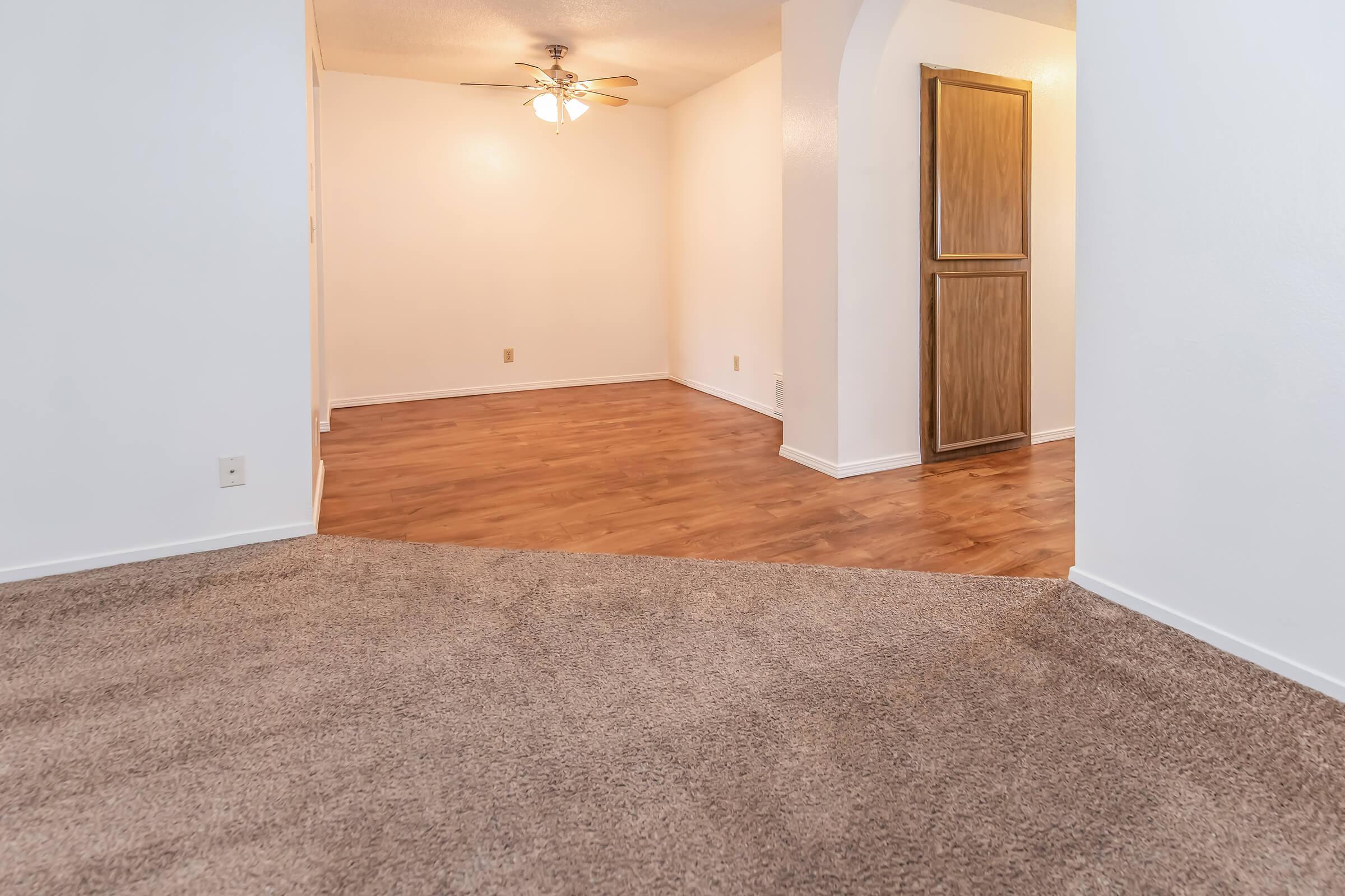 Interior view of a spacious room featuring light-colored walls and a ceiling fan. The flooring transitions from carpet to wood, with a visible archway leading to another area. The lighting gives the space a bright and inviting atmosphere. A wooden cabinet is partially visible on the right.