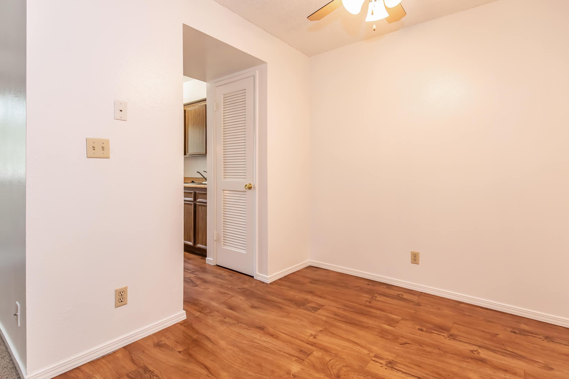 A well-lit room with white walls and a ceiling fan. The floor features wood-like laminate, and there's an open doorway leading to a kitchen area. A closet door is visible on the right. The overall atmosphere is clean and spacious, suitable for various uses like a living area or office.