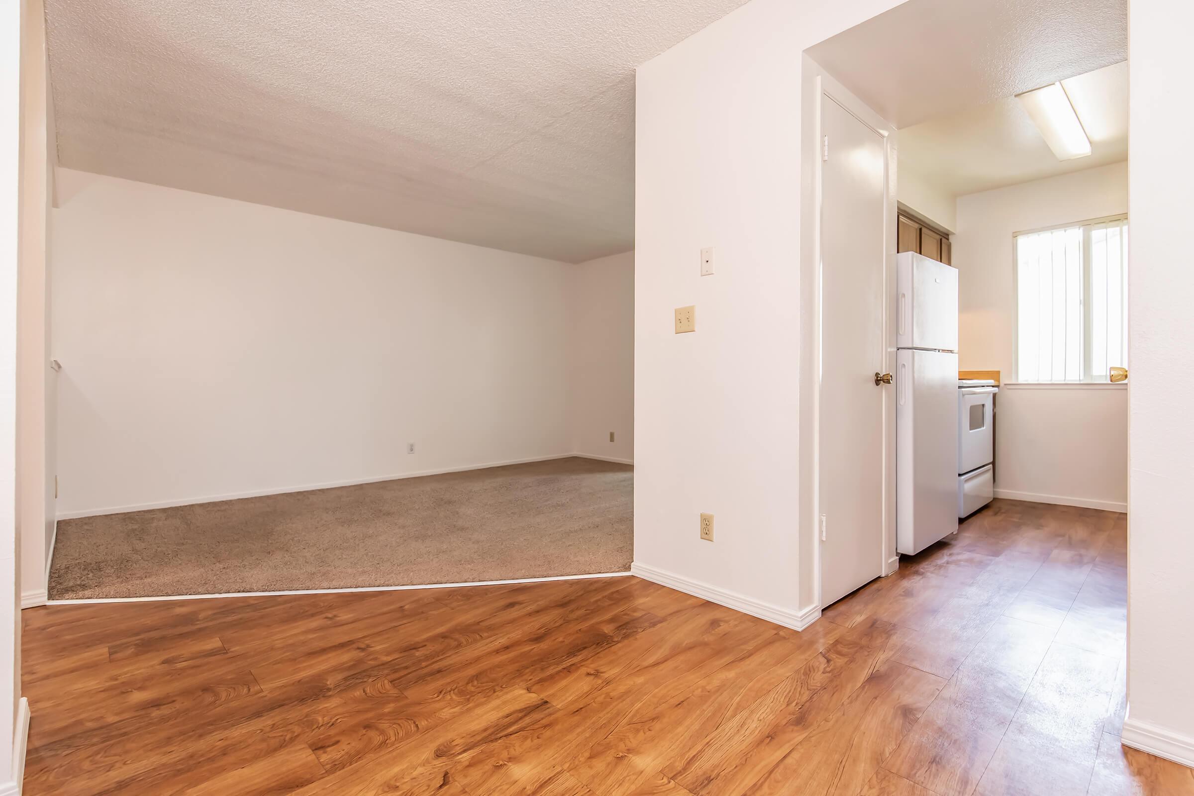 Interior view of a bright, open apartment space. The foreground shows a hardwood floor leading into a carpeted area, while the background features a small kitchen with a white refrigerator and stove. Large windows allow natural light into the room, emphasizing the clean and spacious layout.