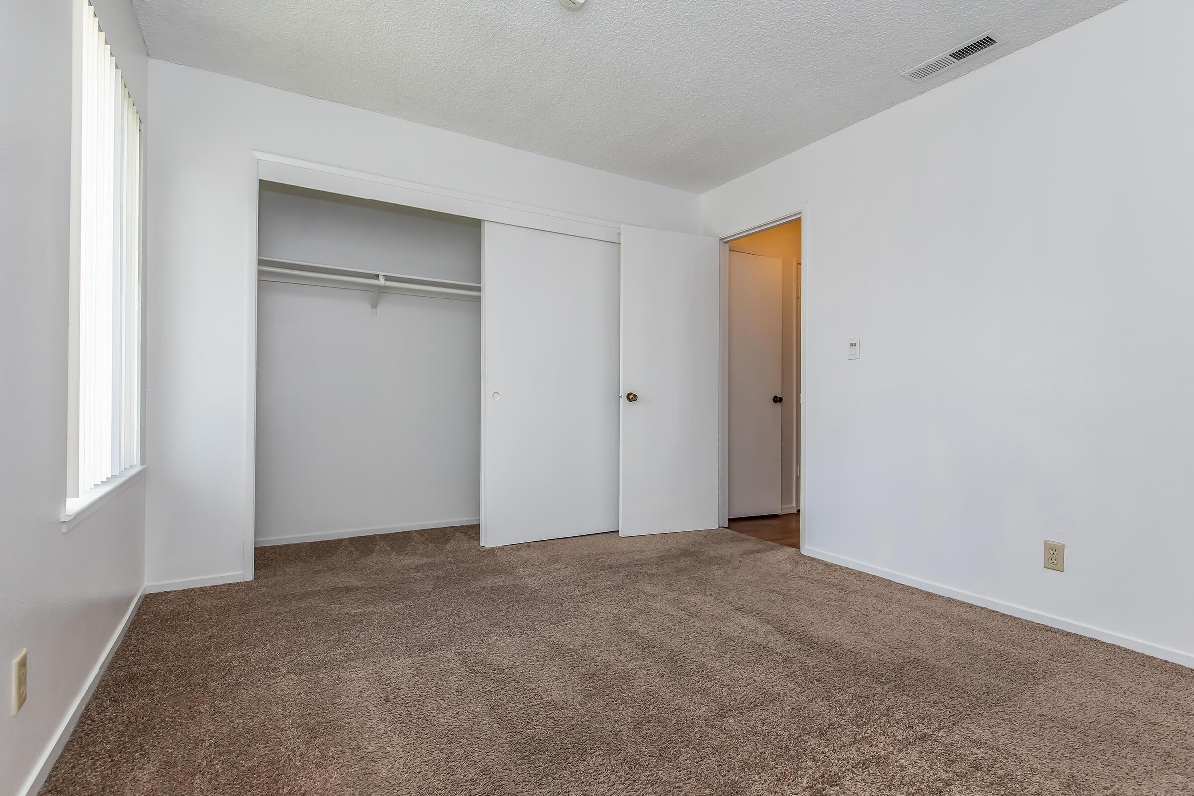 Interior of a simple, empty room with beige carpet flooring. There are white walls with a closet featuring sliding doors on the left, and an open door leading to another space on the right. Natural light enters through a nearby window, and the overall atmosphere is clean and bright.