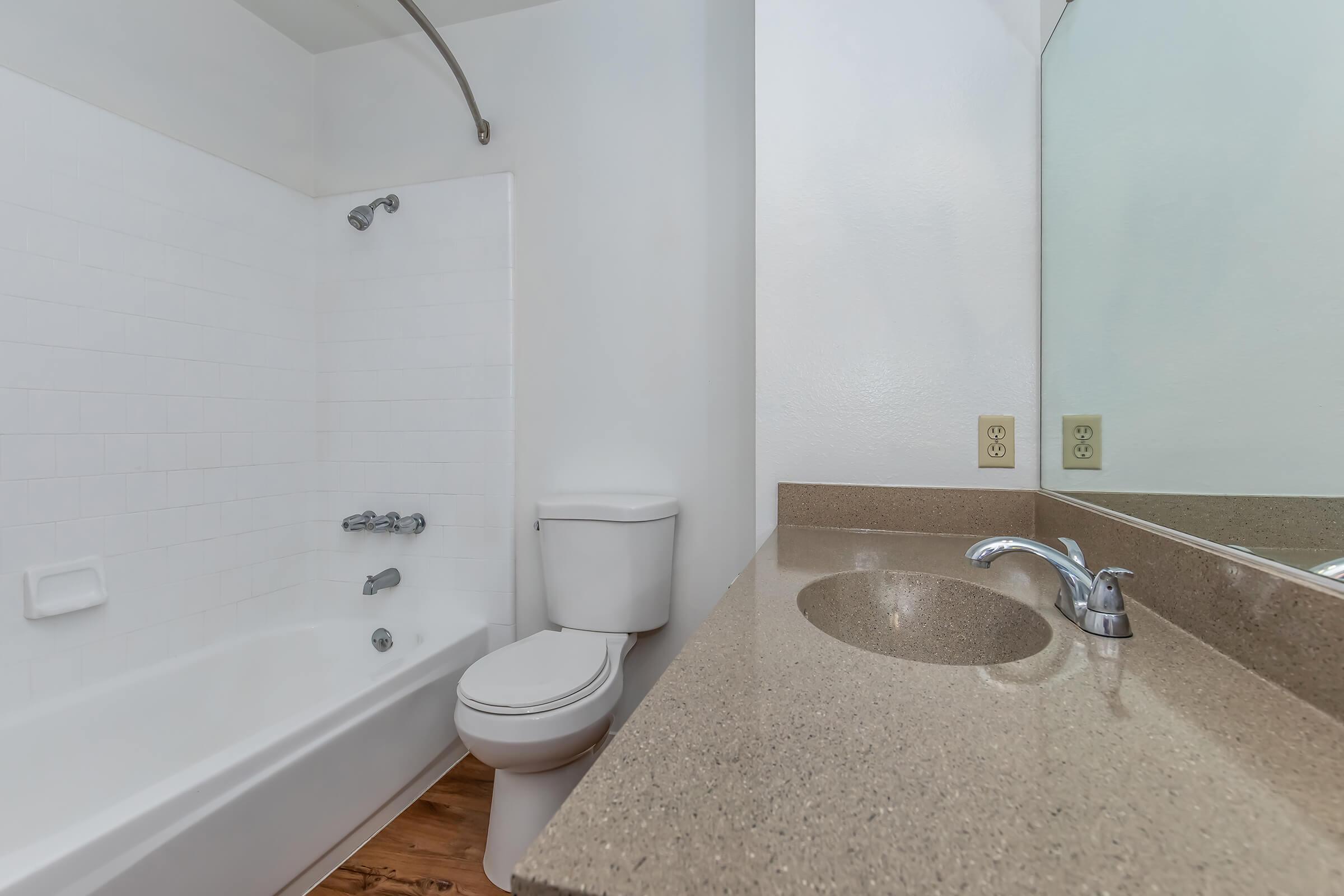 A clean and modern bathroom featuring a white bathtub with shower, a toilet, and a vanity with a smooth countertop and sink. The walls are painted white, and the floor has a wood finish, adding a touch of warmth to the space. A mirror is mounted above the vanity.