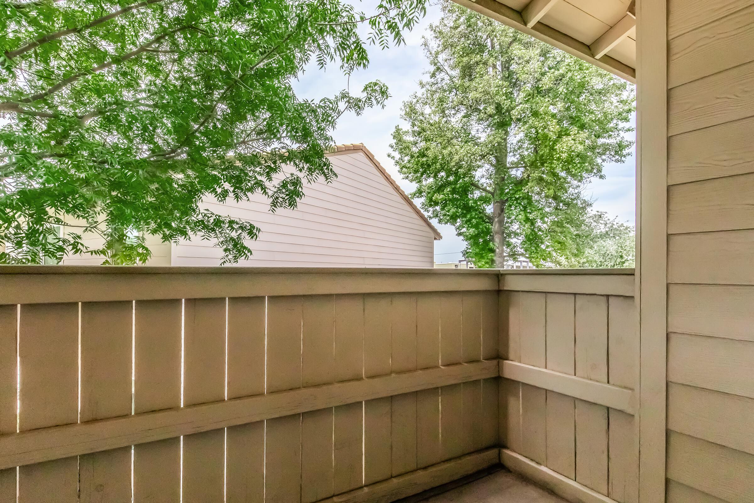A view from a balcony featuring wooden fencing, green leaves from nearby trees, and light-colored houses in the background under a cloudy sky. The scene conveys a peaceful outdoor space.