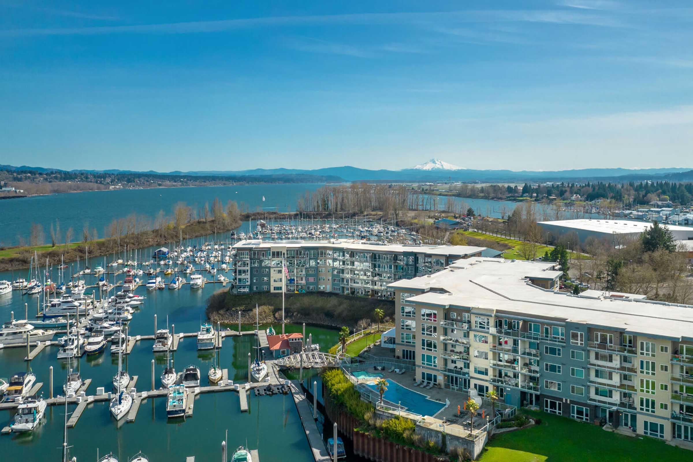 Aerial view of a marina with numerous boats docked. In the foreground, a modern waterfront building is visible, along with a surrounding landscape of trees and hills. In the background, a mountain can be seen under a clear blue sky, creating a serene coastal atmosphere.