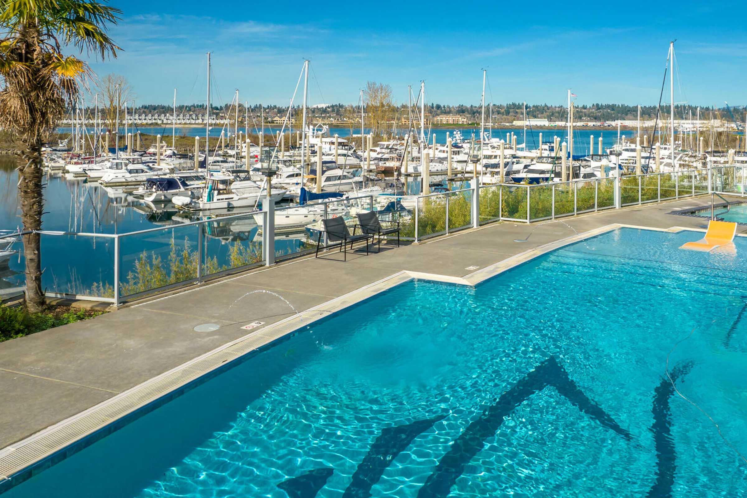 Swimming pool with clear blue water in the foreground, featuring lounge chairs next to it. In the background, there is a marina filled with boats and sailboats, with a landscape of trees and hills under a bright blue sky.