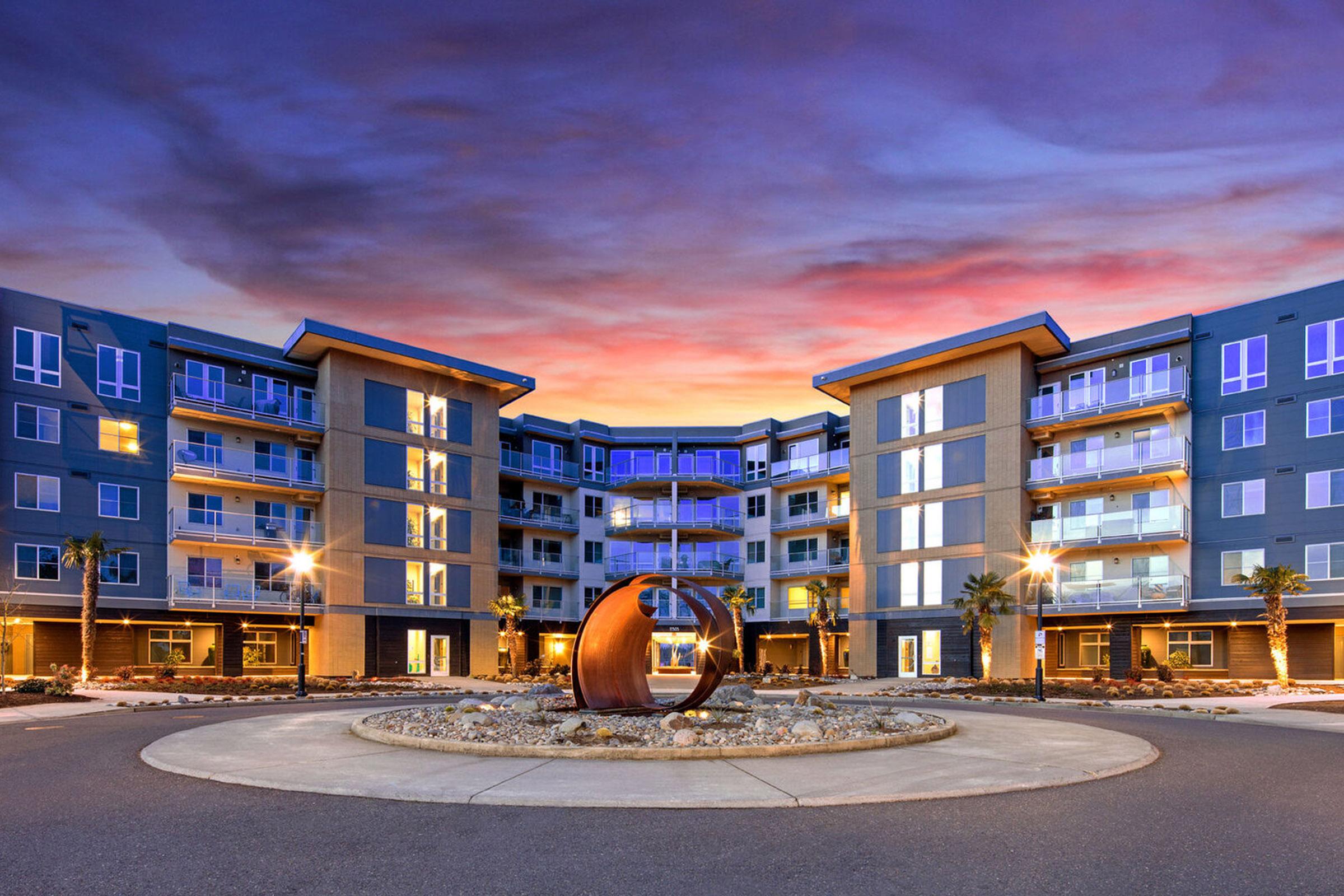 Modern apartment building at twilight, featuring contemporary architecture with multiple floors, large windows, and balconies. A circular driveway with a decorative sculpture and landscaped areas with palm trees surrounds the building, all under a vibrant sky with shades of purple and orange.