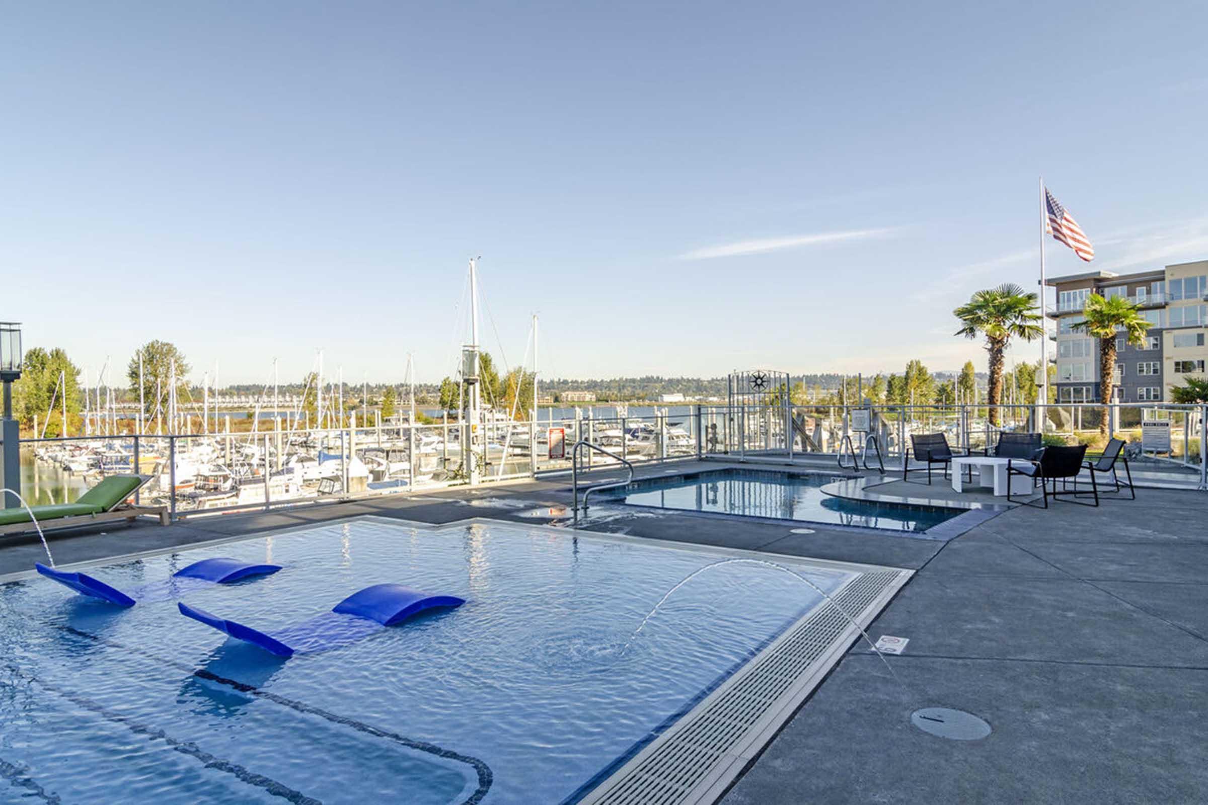 A rooftop pool area featuring blue lounge chairs and a Jacuzzi, overlooking a marina filled with boats. The scene is framed by palm trees and modern buildings, with an American flag waving in the background against a clear blue sky.