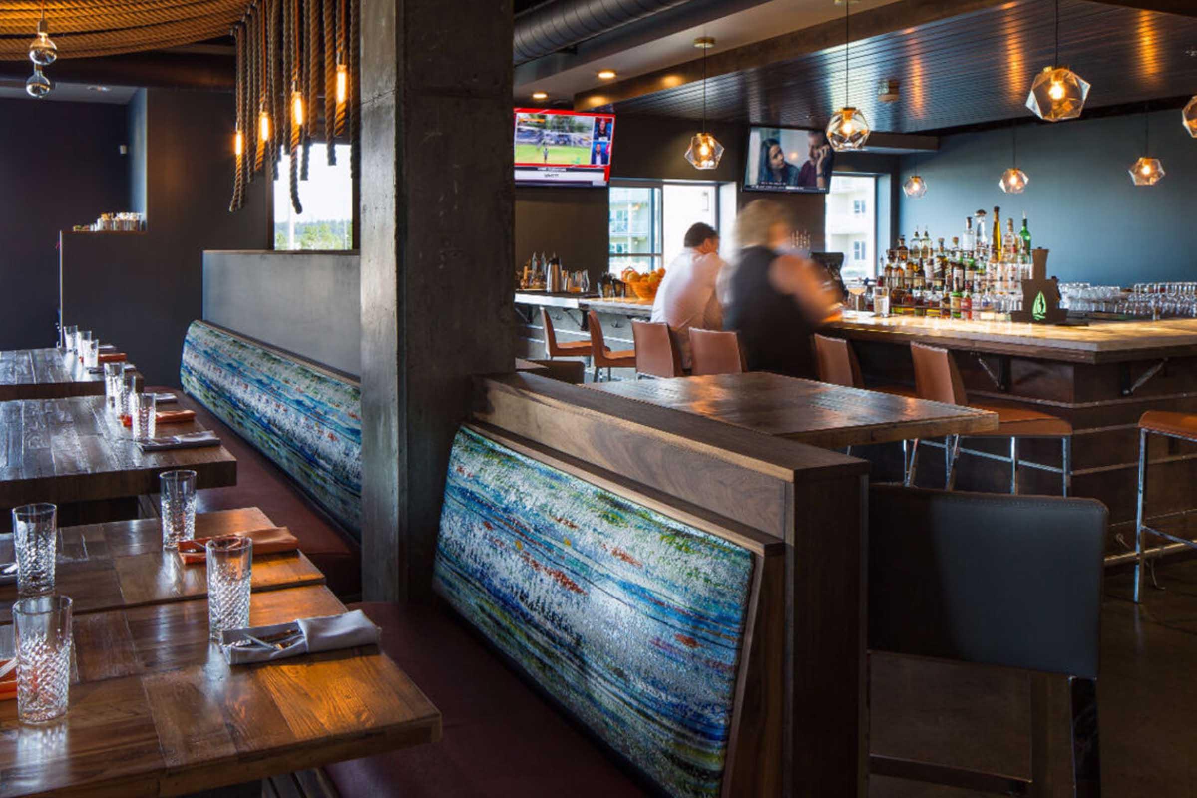 Interior view of a modern restaurant featuring dark wood furnishings, pendant lighting, and a bar area. The tables are set with glassware, and colorful artwork is displayed on the walls. There are televisions mounted on the wall, and staff members can be seen attending to customers.