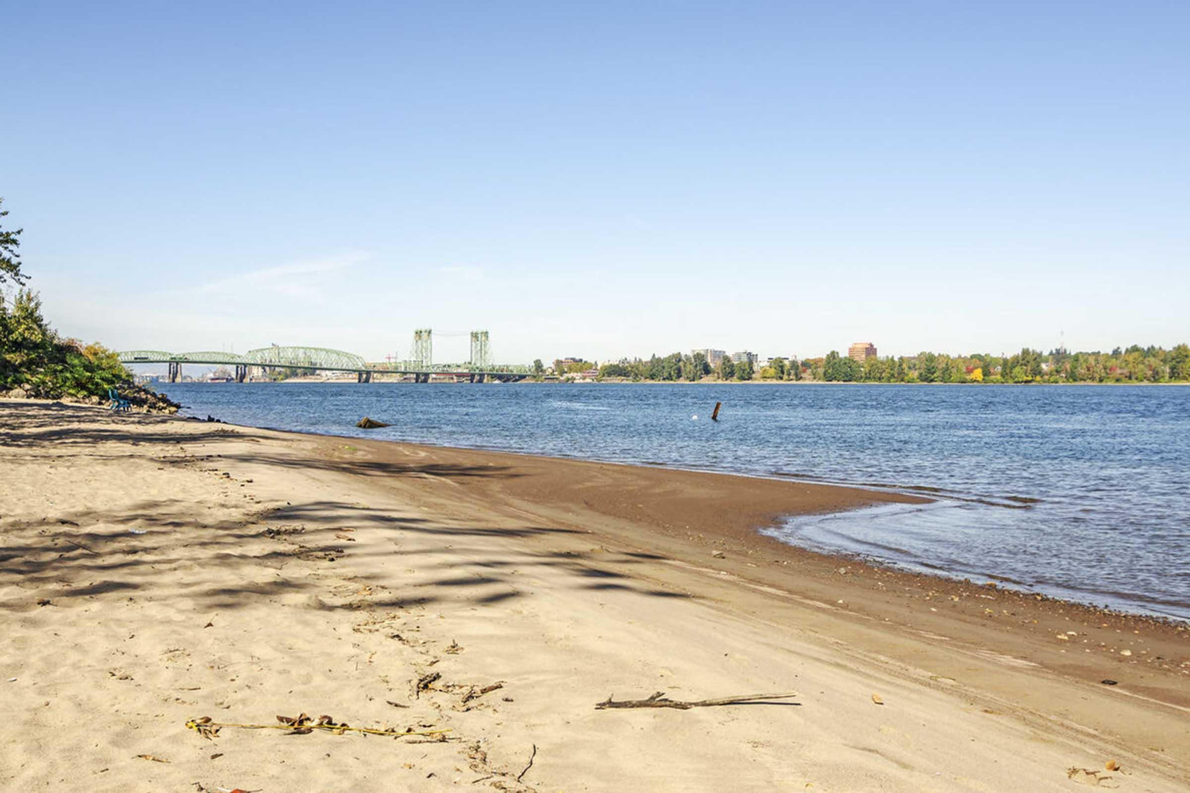 A serene beach scene featuring soft sand, a calm river, and a clear blue sky. In the distance, a green bridge spans the water, surrounded by trees with autumn foliage. The tranquil landscape invites relaxation and enjoyment of nature.