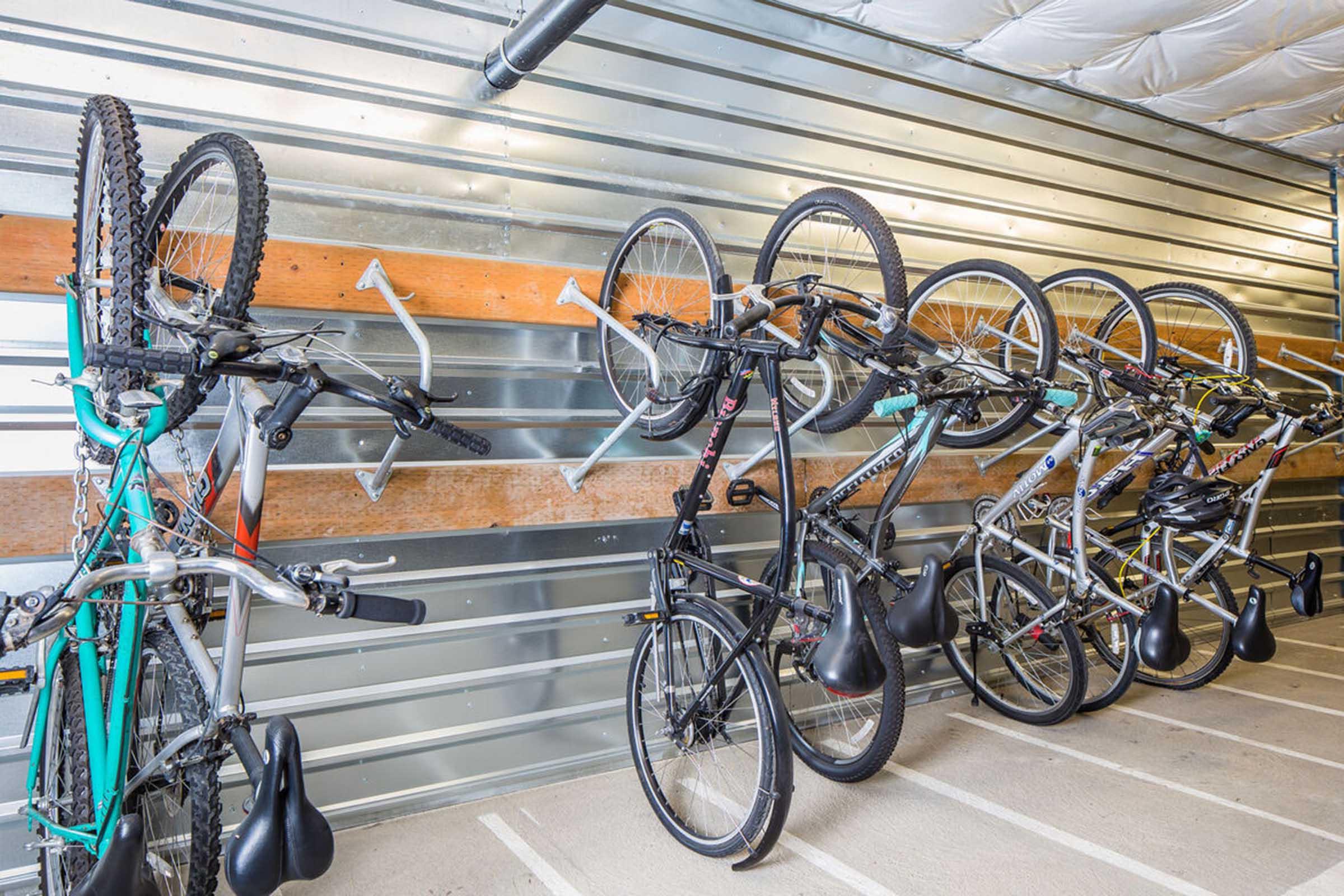 A row of bicycles stored vertically on a metal wall rack in a parking area. The bikes vary in colors and styles, with some upside down and some right side up. The storage area features metal walls and a clean, organized interior.