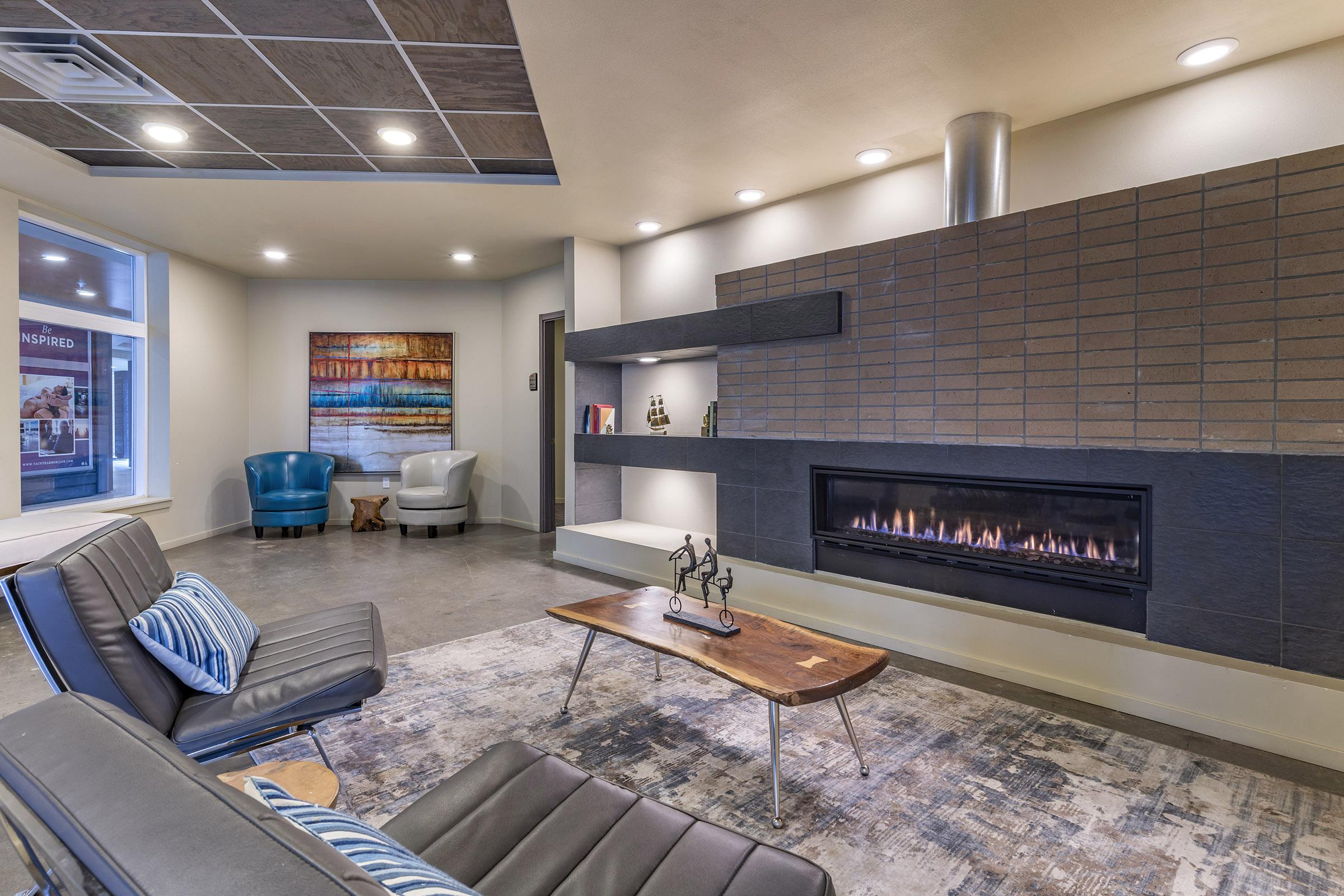 Modern living room with a cozy fireplace, minimalist furniture, and a coffee table. Stylish blue and gray armchairs are present, along with decorative shelves and artwork on the wall. Soft lighting creates a warm ambiance in the space.