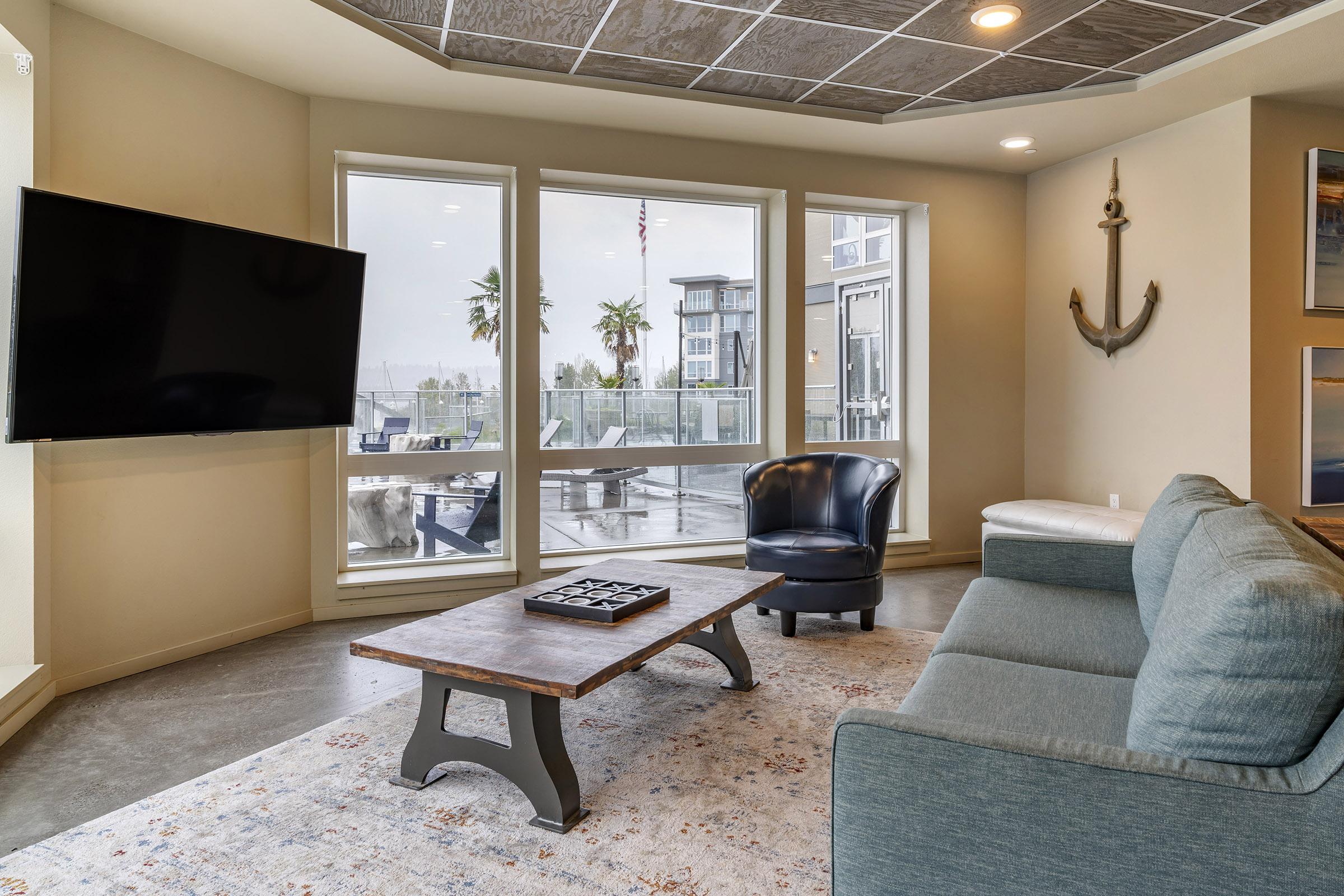 A modern living room featuring a blue sofa, a black armchair, and a wooden coffee table with a decorative item. Large windows let in natural light, showcasing an outdoor area with palm trees and a glimpse of water. The space has a contemporary aesthetic with a flat-screen TV mounted on the wall.