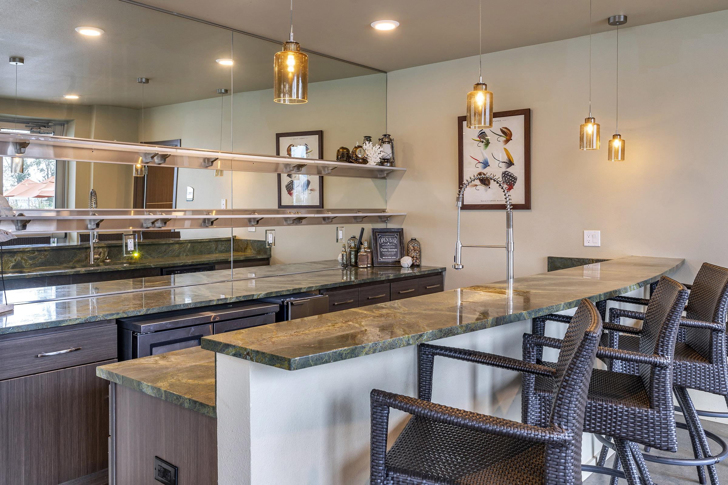 Modern kitchen with a long green stone countertop, featuring stylish bar stools. The space includes a mirrored backsplash, decorative items on the shelves, and overhead pendant lights, creating a warm and inviting atmosphere. A framed artwork of birds adds a decorative touch to the wall.