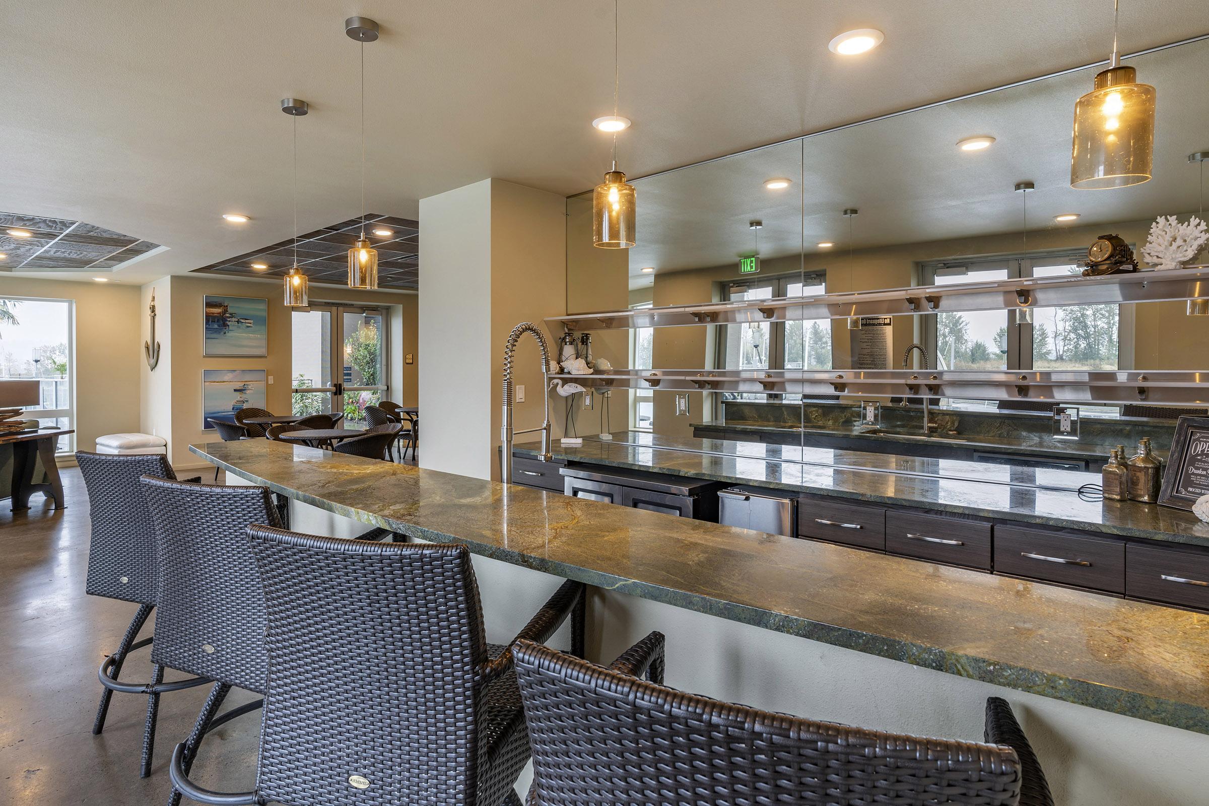 A modern, stylish kitchen with a long countertop featuring four black wicker bar stools. Above the counter, pendant lights hang from the ceiling. The space has a minimalist design with a large mirror reflecting the room and a view of outdoor greenery through large windows.