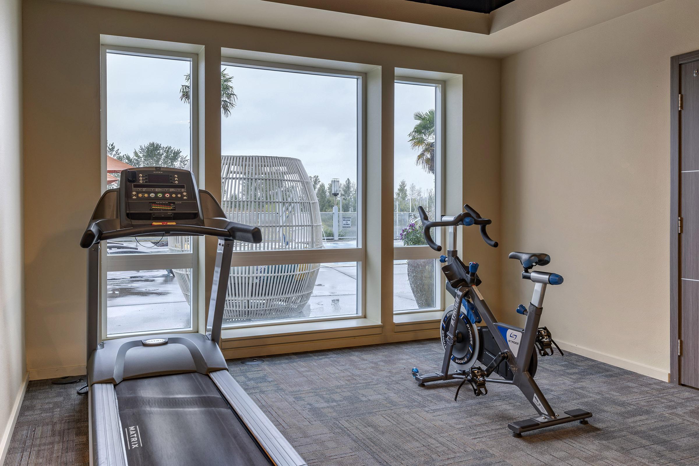 A fitness room featuring a treadmill and a stationary bike, with large windows providing a view of outdoor greenery. The light-colored walls and carpet create a spacious ambiance, enhancing the workout environment.