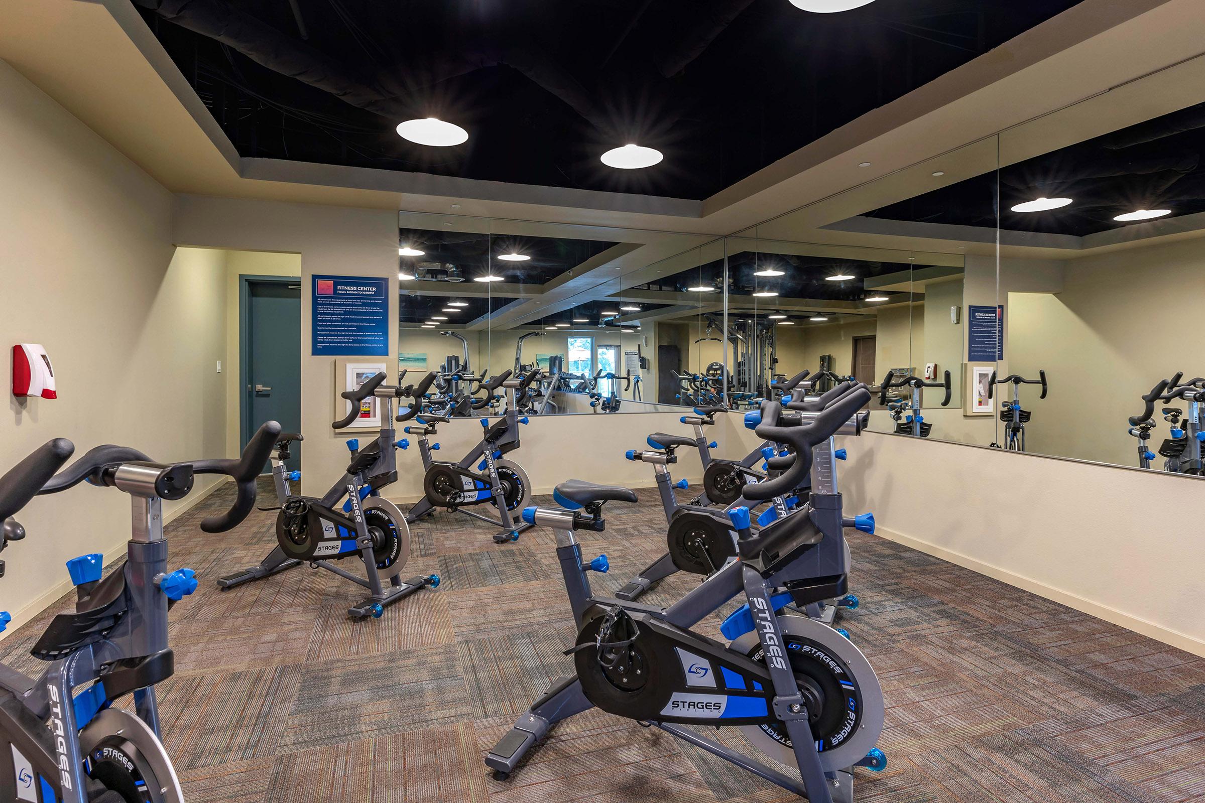 A modern indoor cycling studio featuring several stationary exercise bikes arranged in rows. The room has a large mirror on one wall, bright overhead lights, and a neutral color scheme, creating a vibrant workout environment.
