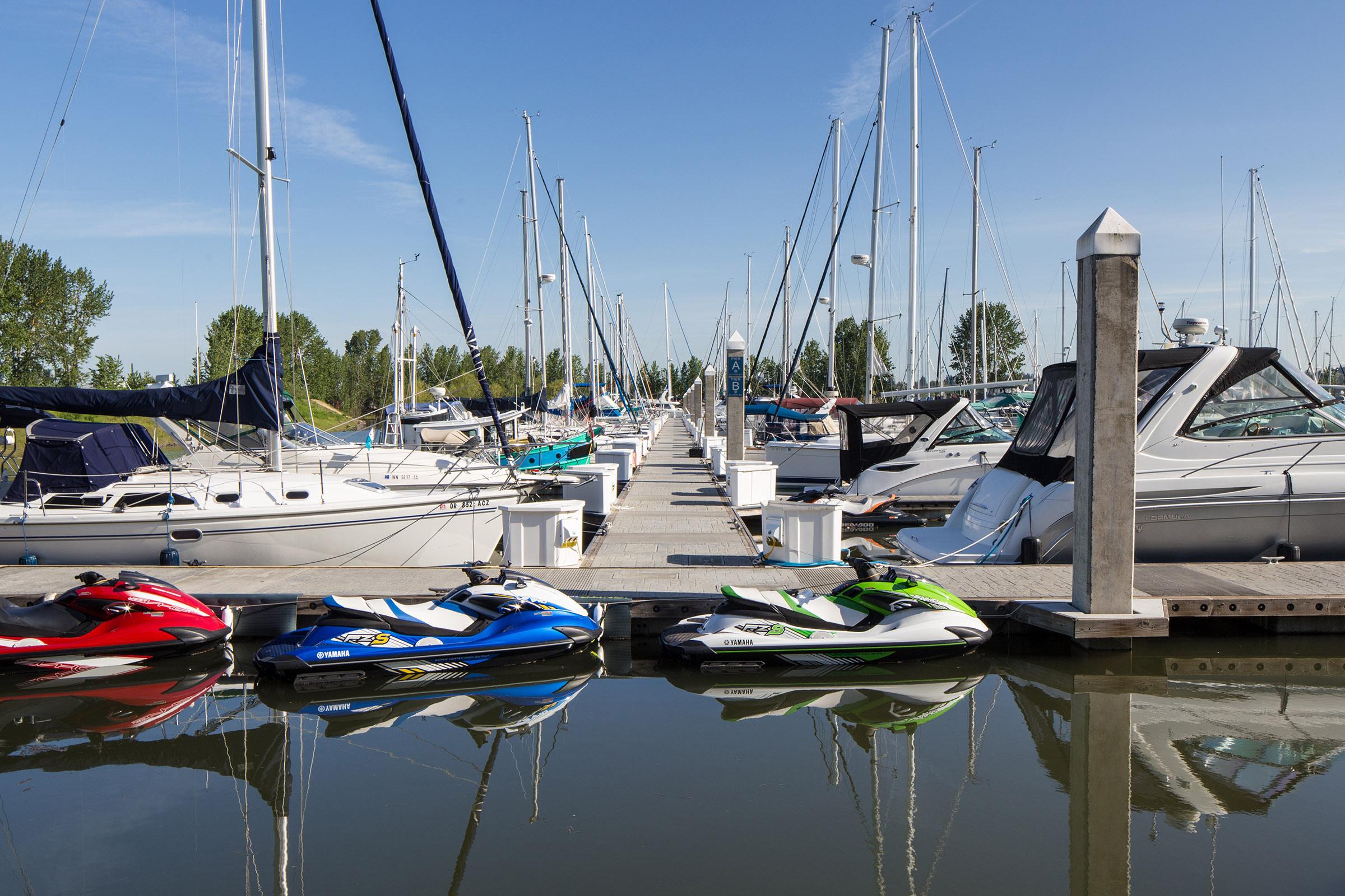 A scenic marina featuring various boats, including sailboats and yachts, lined up along a wooden dock. Colorful jet skis in the foreground reflect in the calm water. Green trees and a clear blue sky complete the picturesque waterfront view.