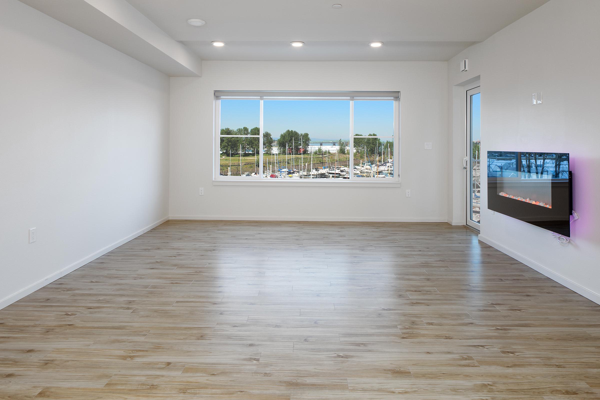 A spacious, empty room with light-colored wooden flooring and large windows allowing natural light. The view outside includes greenery and parked vehicles. There’s a modern wall-mounted fireplace on one side, adding a cozy element to the minimalist decor.