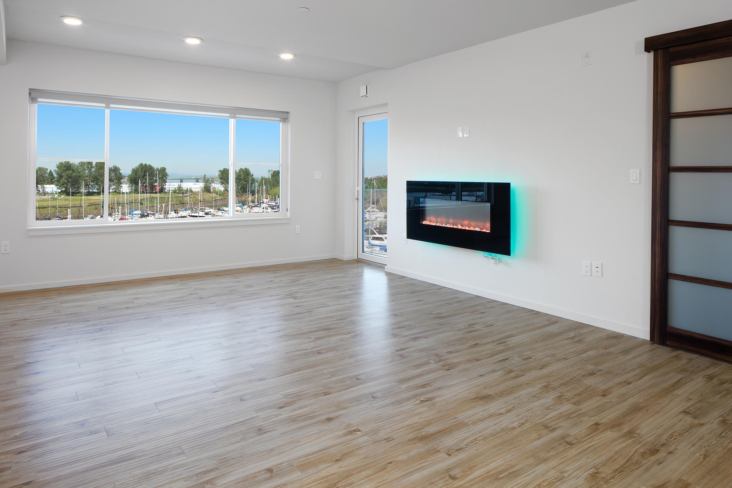 A modern living room featuring large windows with a view of greenery and a marina. The space has light wood flooring, a white wall with an electric fireplace mounted on it, and a glass door leading outside. Bright, natural light fills the room, creating an inviting atmosphere.